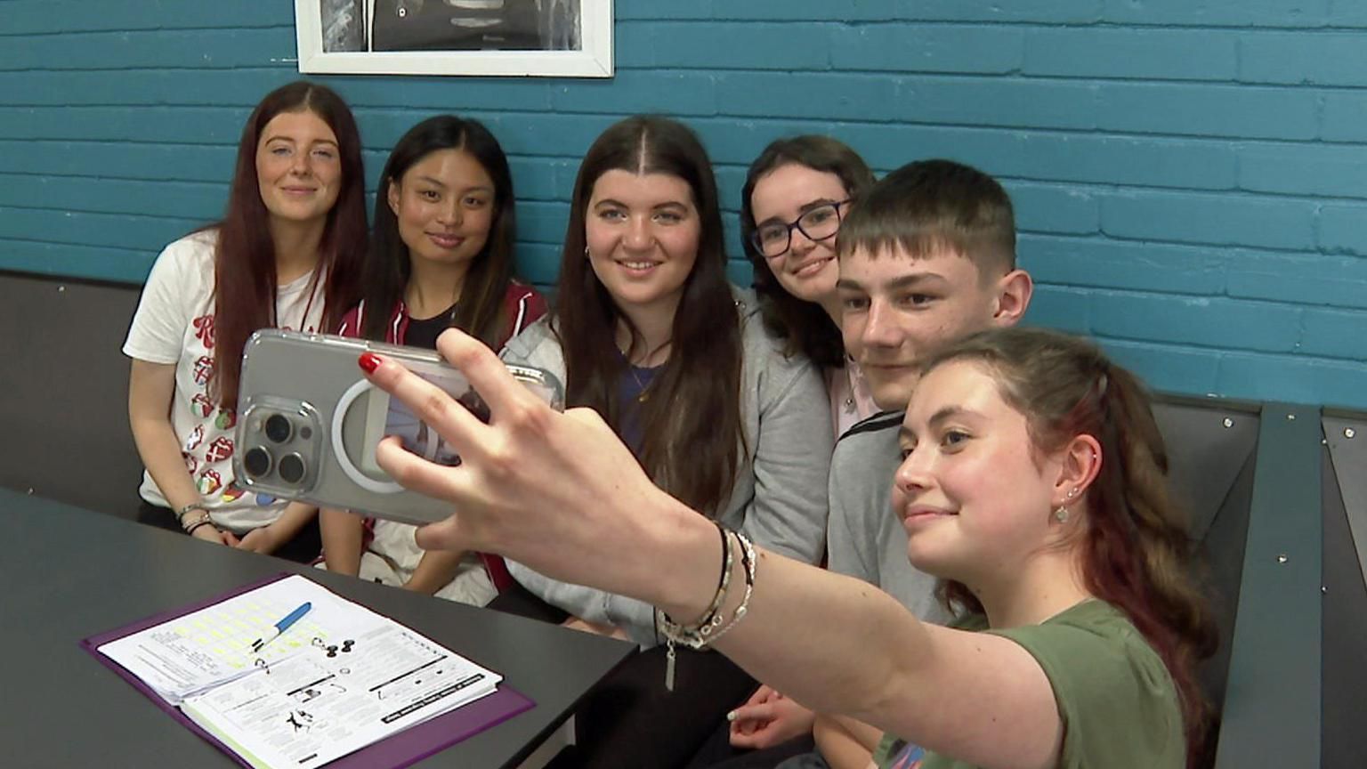 A group of six teenagers are sitting in a row. One is holding up a phone and taking a selfie of the whole group.