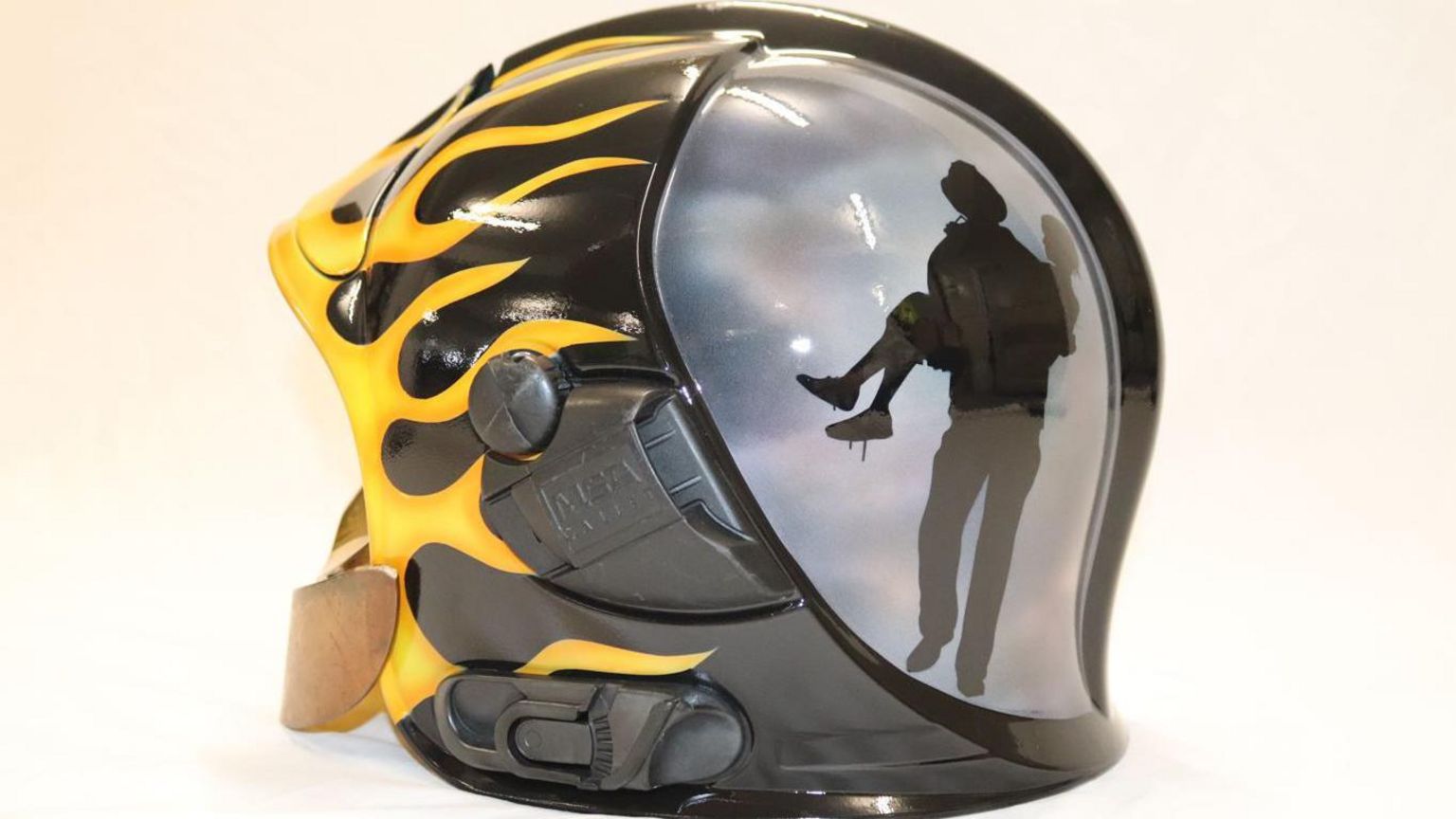 The rear side of a firefighters helmet painted in black with yellow and orange hot rod flames and a silhouetted image of a firefighter rescuing  a girl