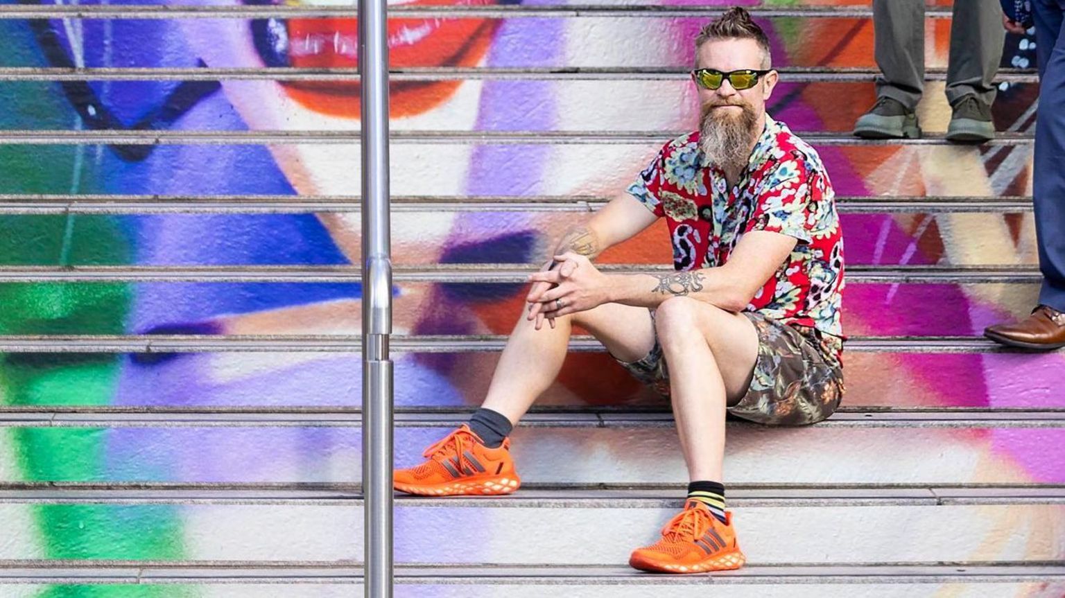 Frank Styles sitting on the steps, which are covered in a Taylor Swift image. He has a beard and wears sunglasses, and is dressed in shorts and a shirt, with orange trainers