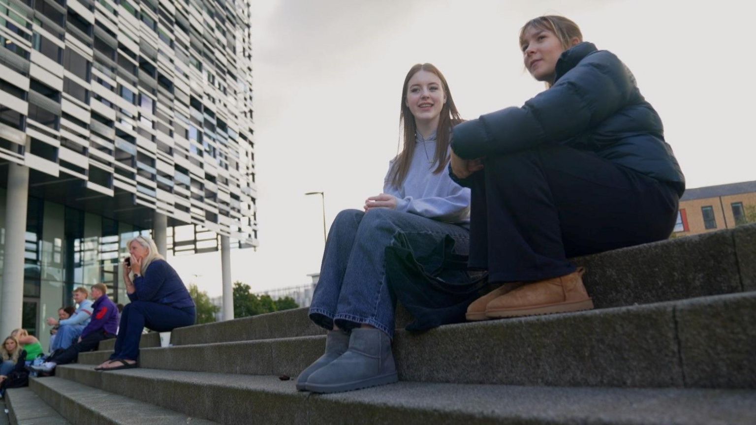 Two students sit on concrete steps. One has long brown hair and a light blue top and jeans. The second is wearing a black shiny jacket. In the background there are other students sitting down. 