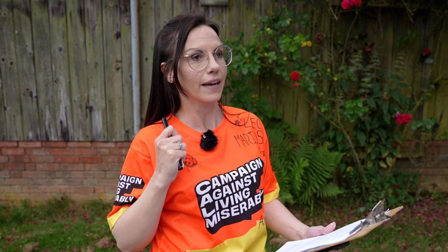 Naomi Woodford holding a clipboard and pen, and speaking into a microphone that is attached to her orange T-shirt. Her T-shirt has "Campaign Against Living Miserably" across its front. 