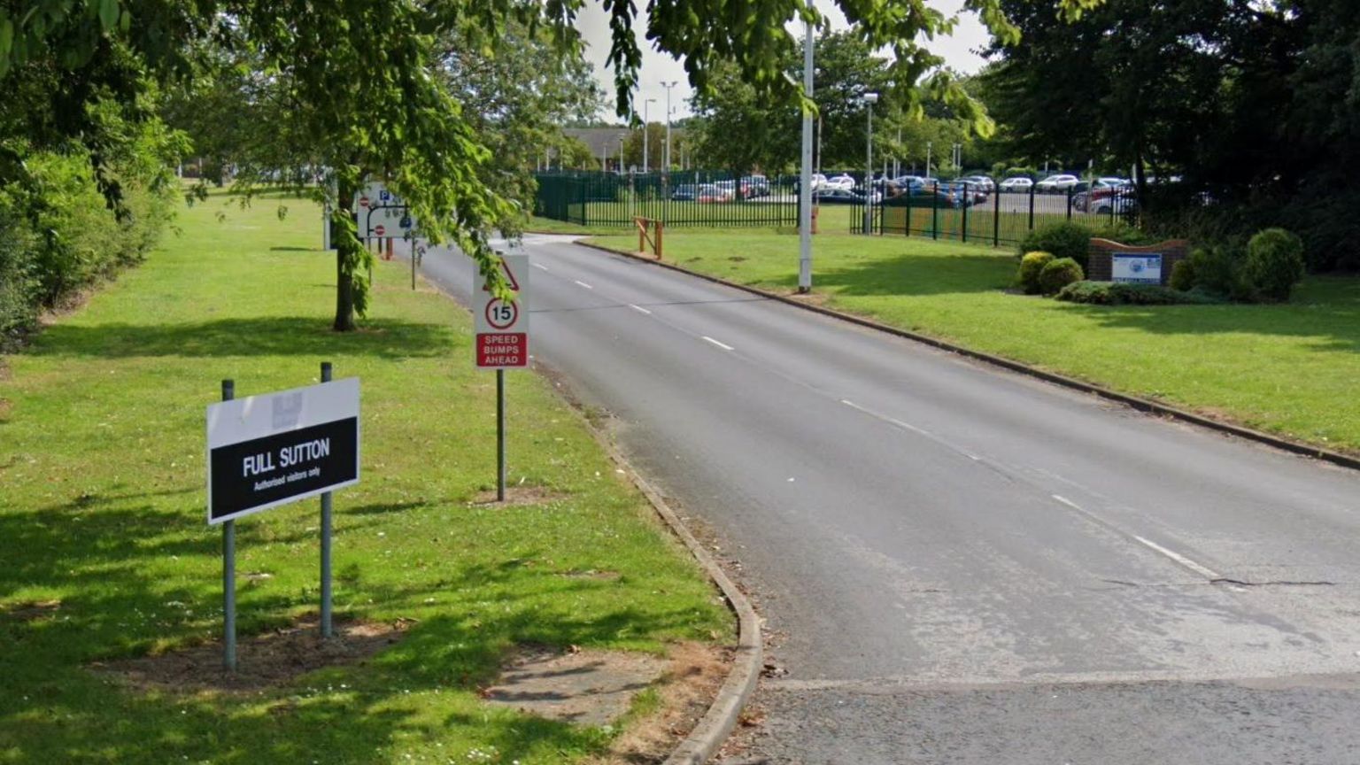 The entrance to HMP Full Sutton. A black and white sign stands on a neatly cut grass verge beside a roadway, with fencing and a car park in the background.