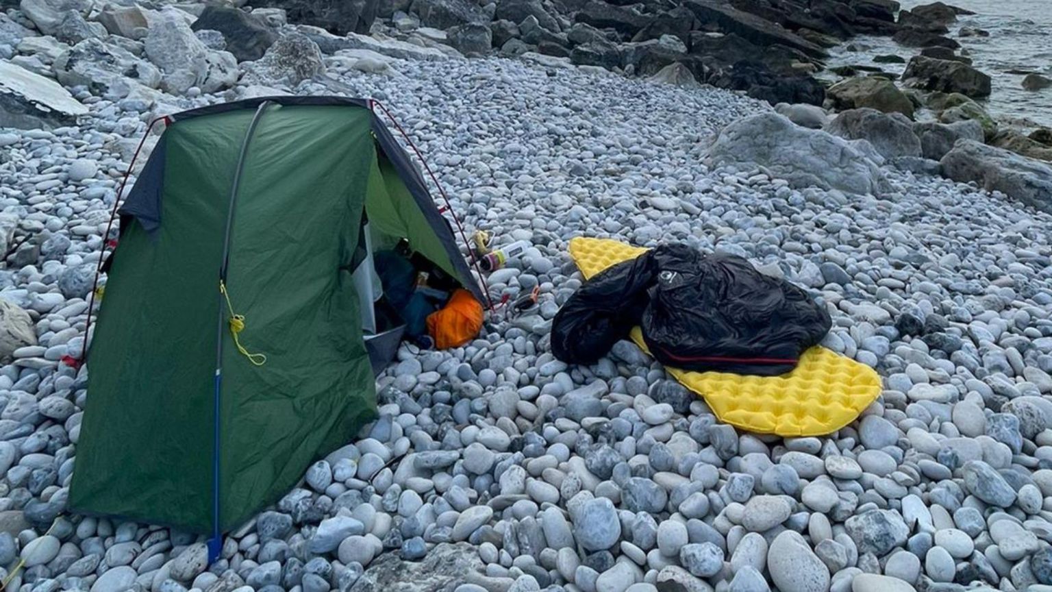 A green tent next to a yellow roll-up camping bed mat laid out on grey pebbles on a beach