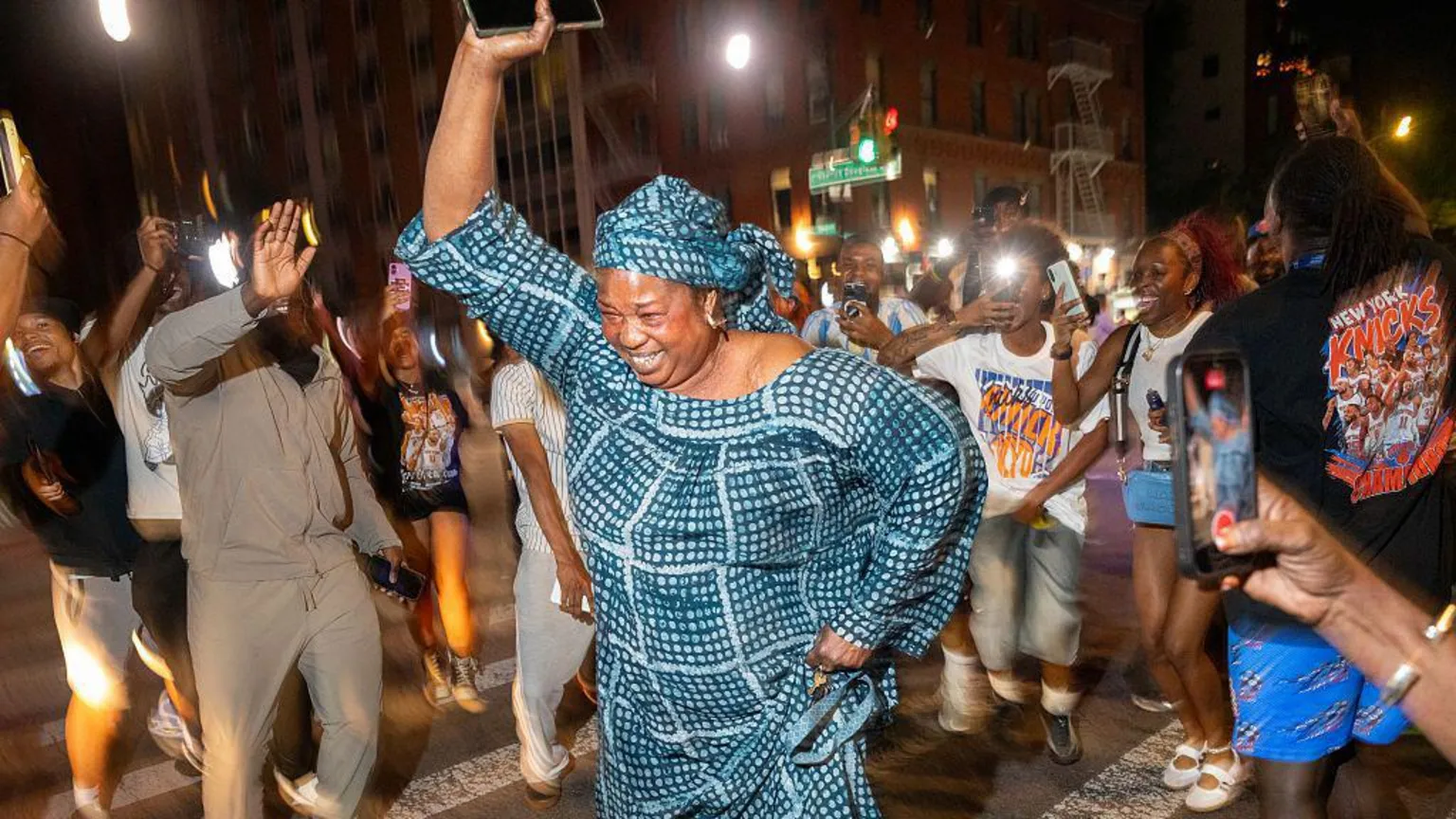 Getty Images A woman wearing a matching headscarf and dress raises her arm in the air as she dances on a zebra crossing in the street at night time. A crowd of other people dance around her, some rais