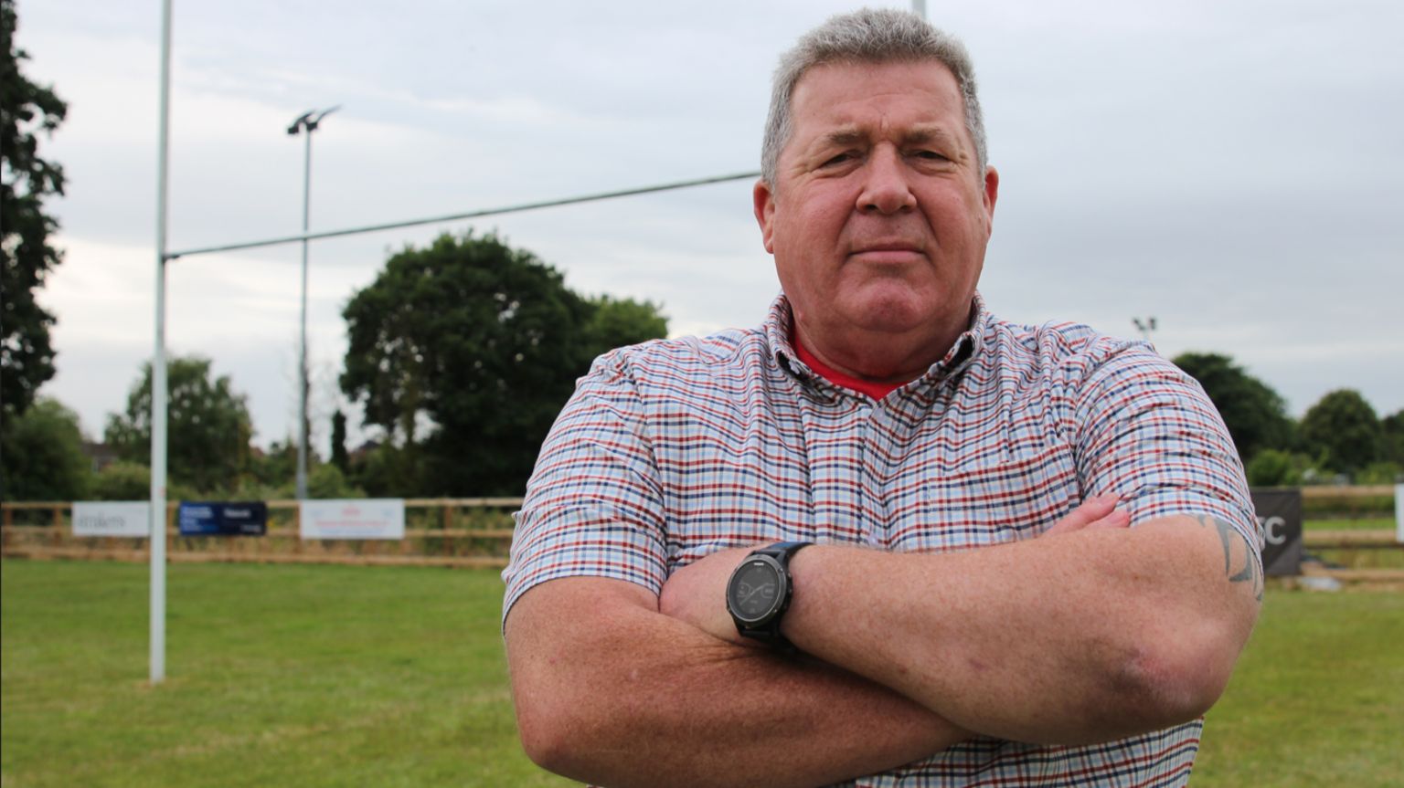 Man in blue and red checked shirt and black watch on his right wrist has his arms folded. There is a rugby post and trees in the background.