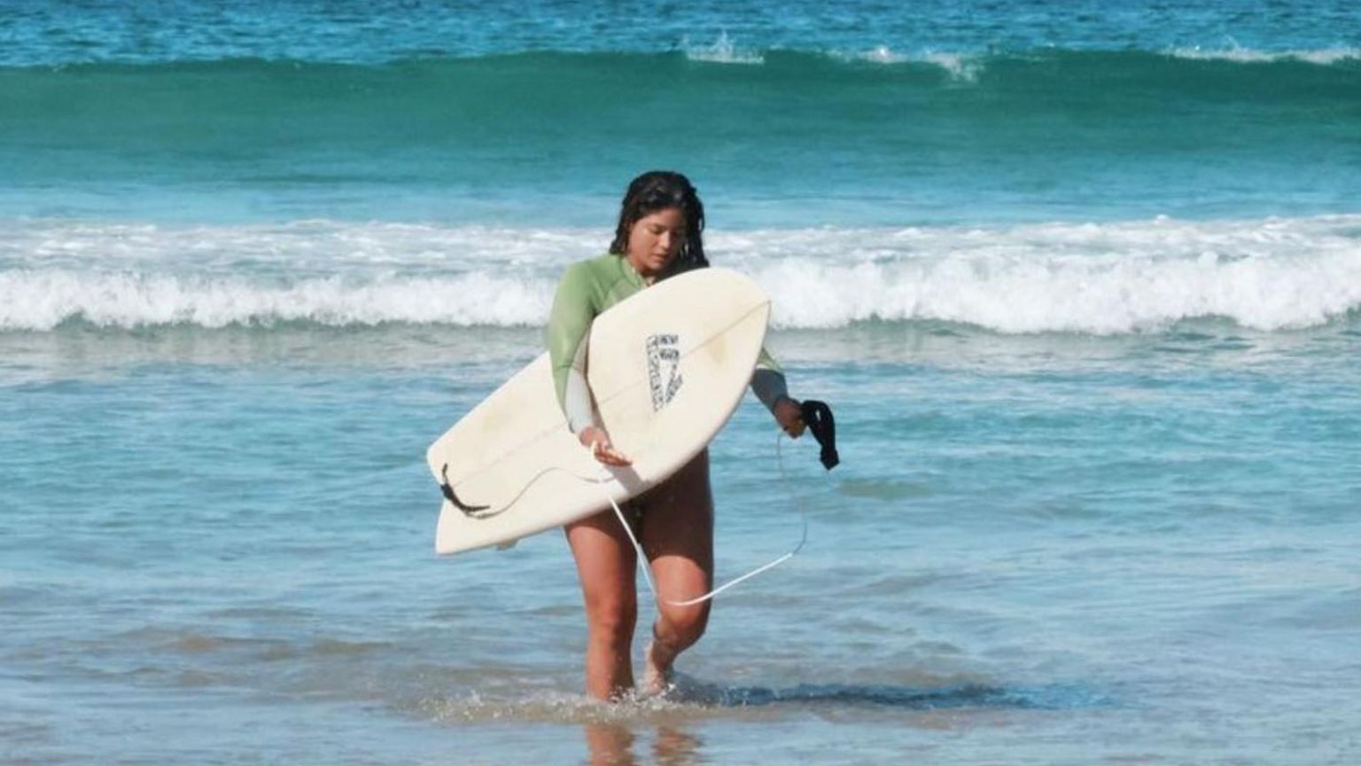 Surfer Mika Flower walking in sea holding a surfboard.