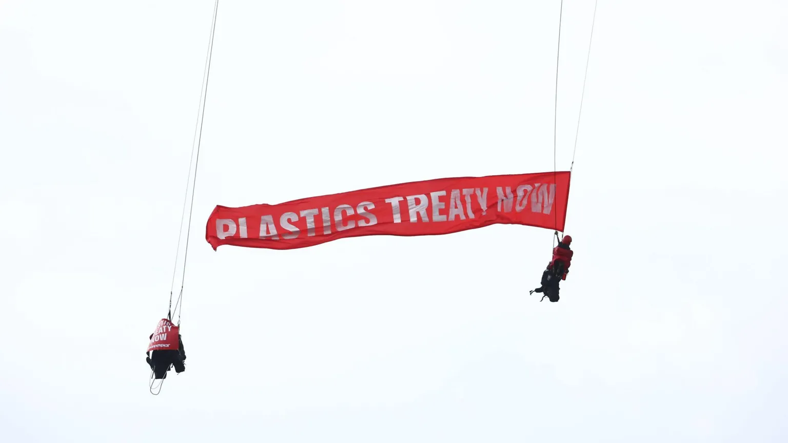The image shows two people suspended from ropes against a white sky, holding a large red banner that reads 