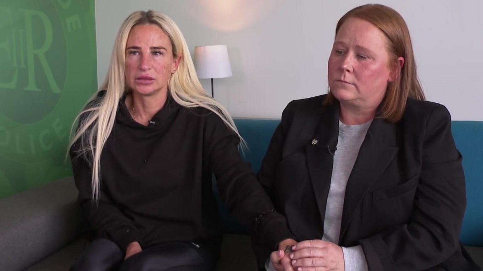 Ellis Cox's mother Carolyn (L) and aunt, Julie O'Toole (R) sit holding each other's hands as they look off from the camera. A green Merseyside Police badge is next to the sofa they're sitting on.

