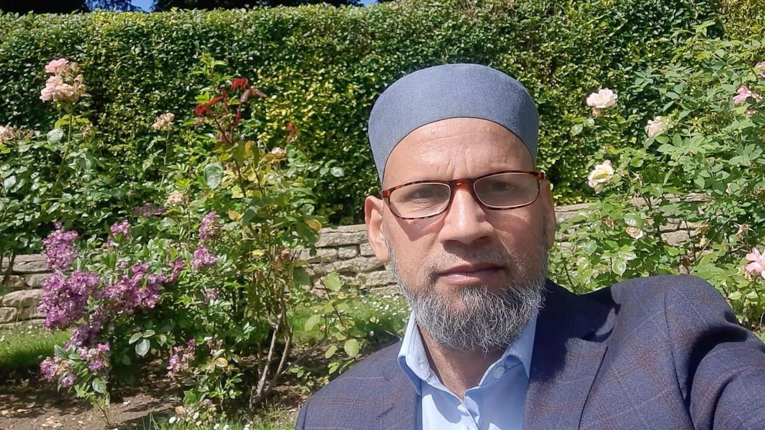 Muhammed Rashid takes a selfie in a garden, he is wearing a blue plaid jacket, blue shirt and kufi