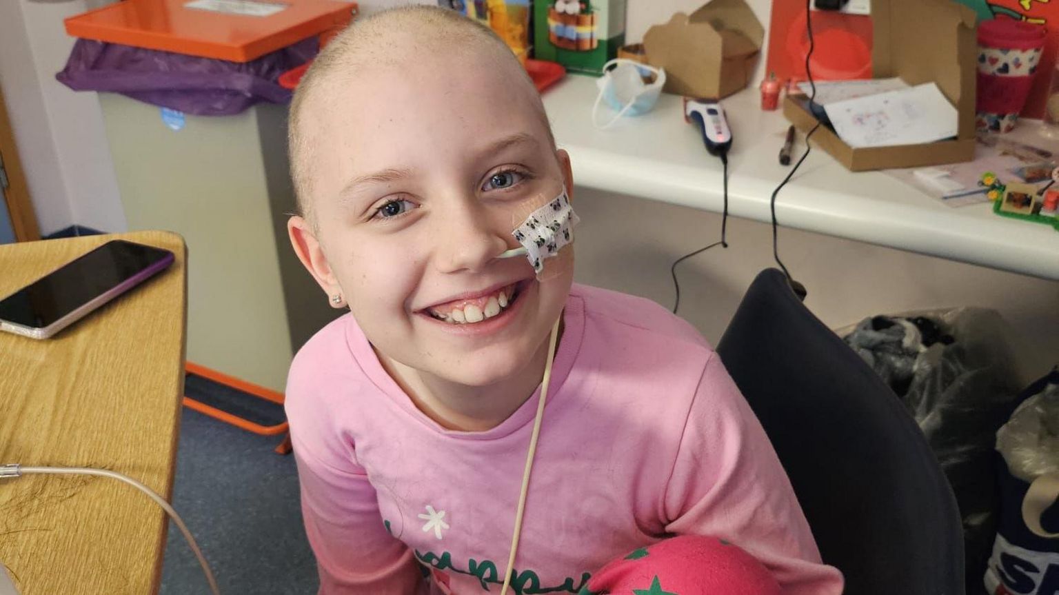 Megan. She is pictured smiling at the camera. She has a small amount of blonde hair and blue eyes. She is wearing a pink t-shirt. 