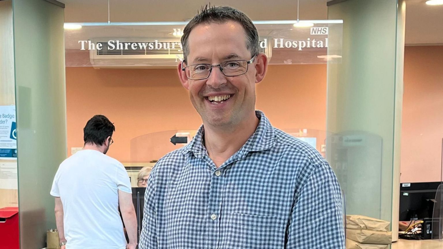 Ed Rysdale, smiling and wearing a blue and white checked shirt and glasses. He is standing in the hospital foyer.