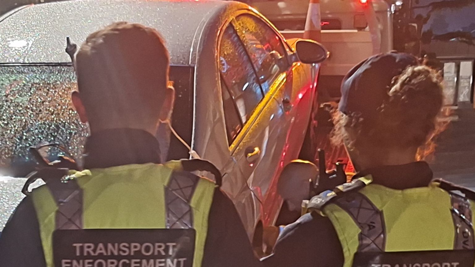 Two transport enforcement officers, one male and one female who is wearing a cap,  stand with their back to camera while looking at a sized grey car being towed away. They are wearing high viz jackets and it is dark