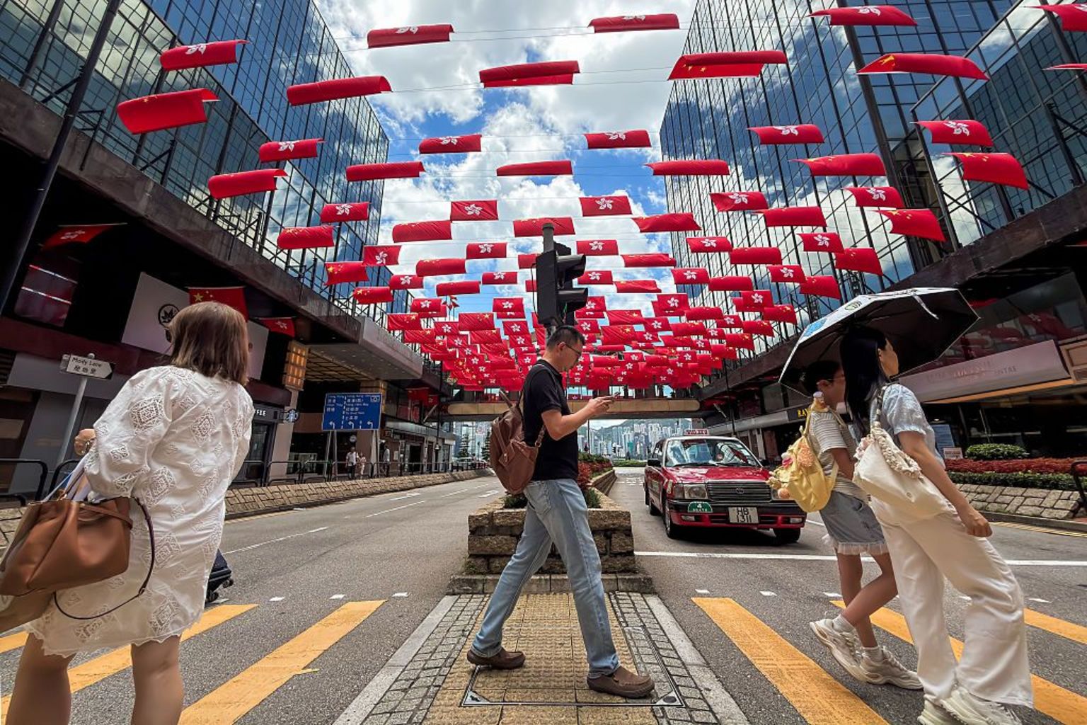 香港街头，行人走过街道，街道两旁办公大楼内悬挂着成排的中国国旗和香港特别行政区旗帜。