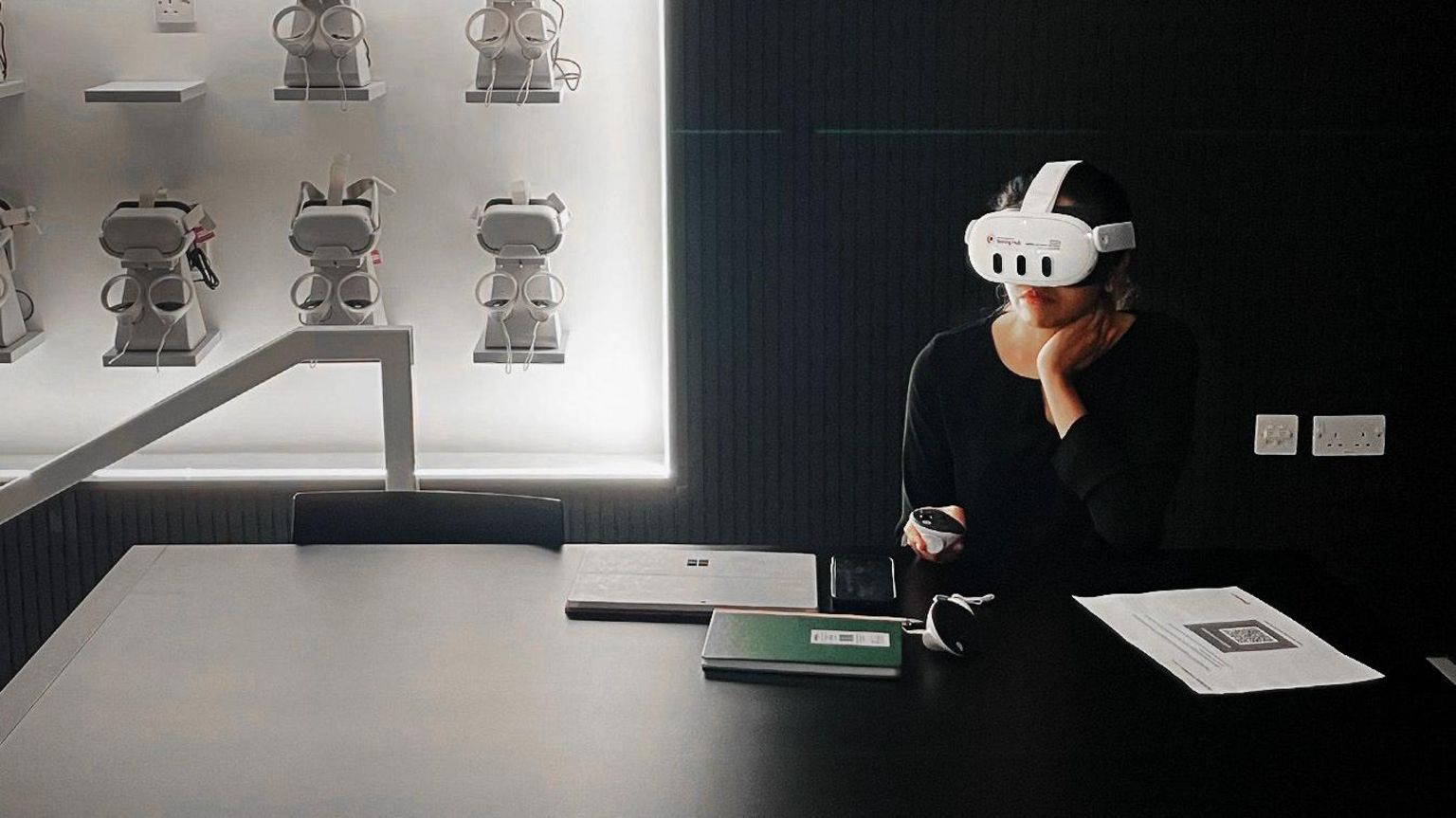 A woman in a black room wearing a VR headset while sitting at a table. 