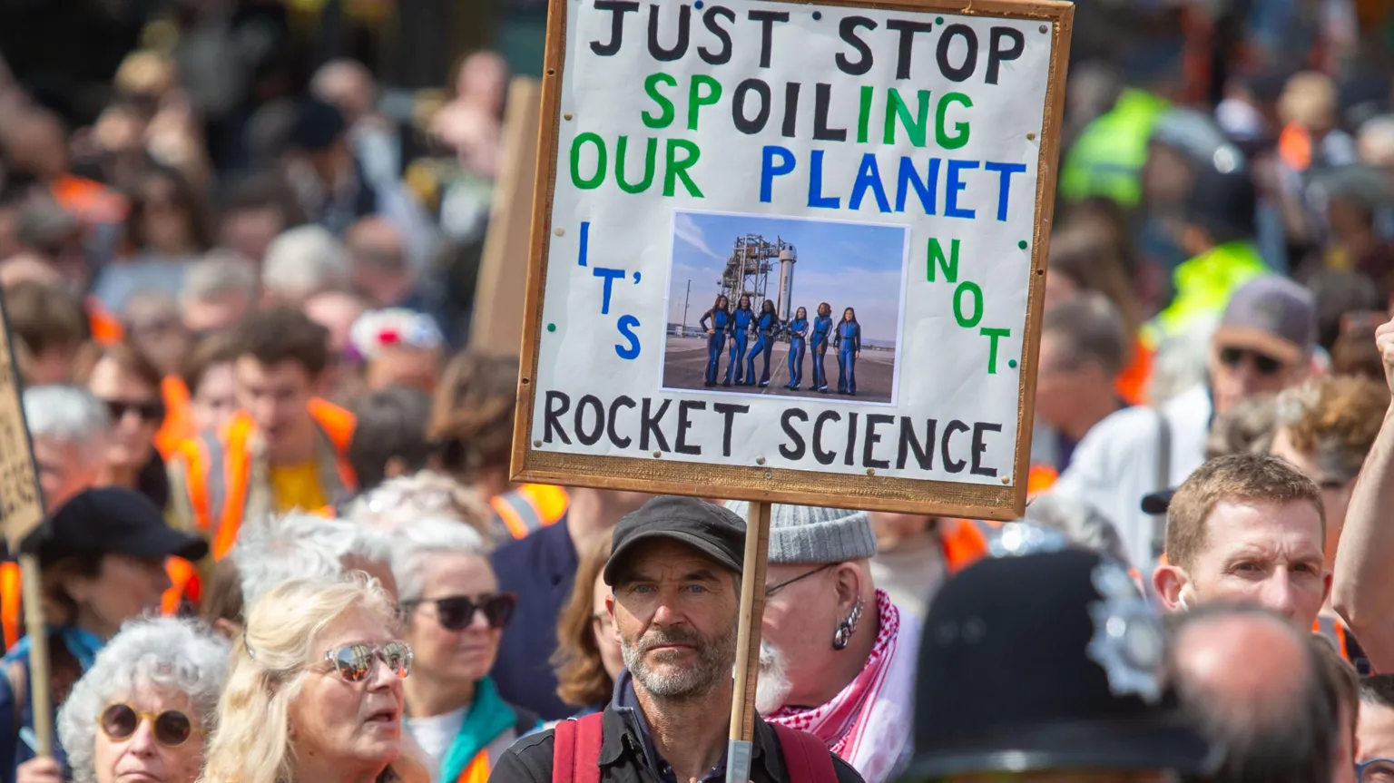The image shows a crowded protest with a prominent sign reading 