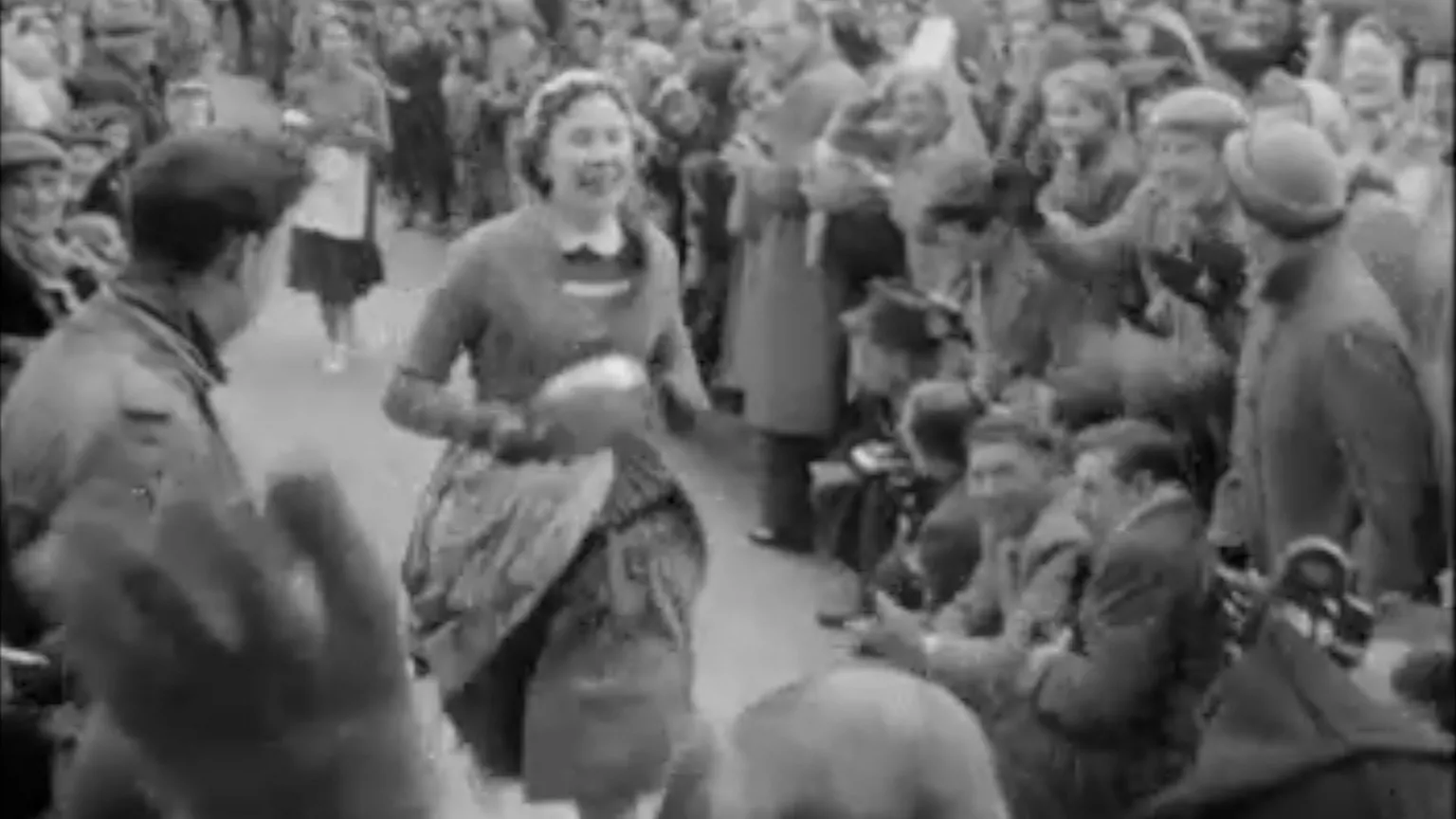 A woman holding a pan run towards the finish line. She is surrounded by onlookers and photographers. The picture is in black and white.