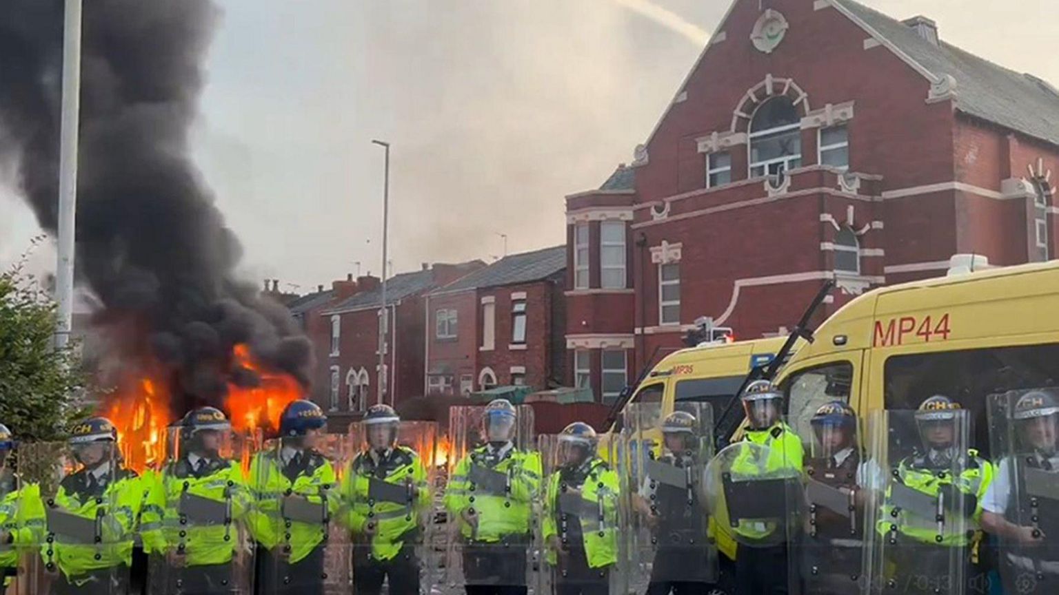 A row of riot police stood in front of two police riot vans outside a mosque where a vehicle is on fire