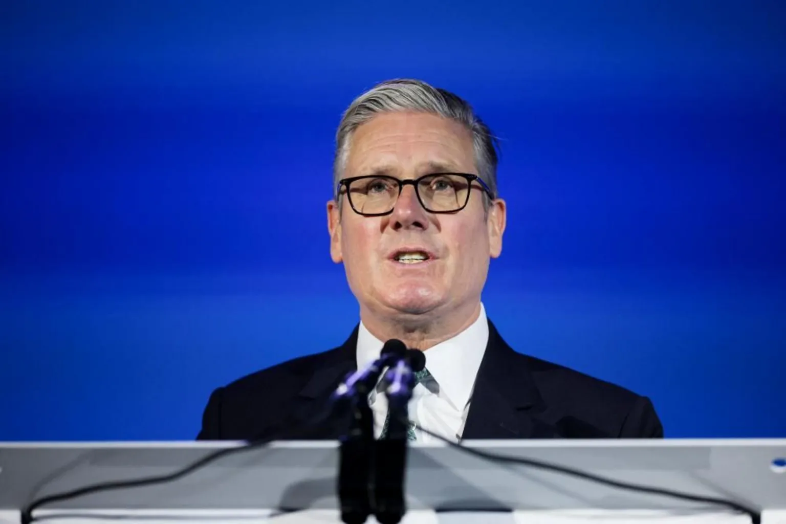 PA Media The prime minister dressed in a dark suit, white shirt and patterned tie is sign behind a podium adorned with two microphones pointing at him. The background is a sheer blue sheeting