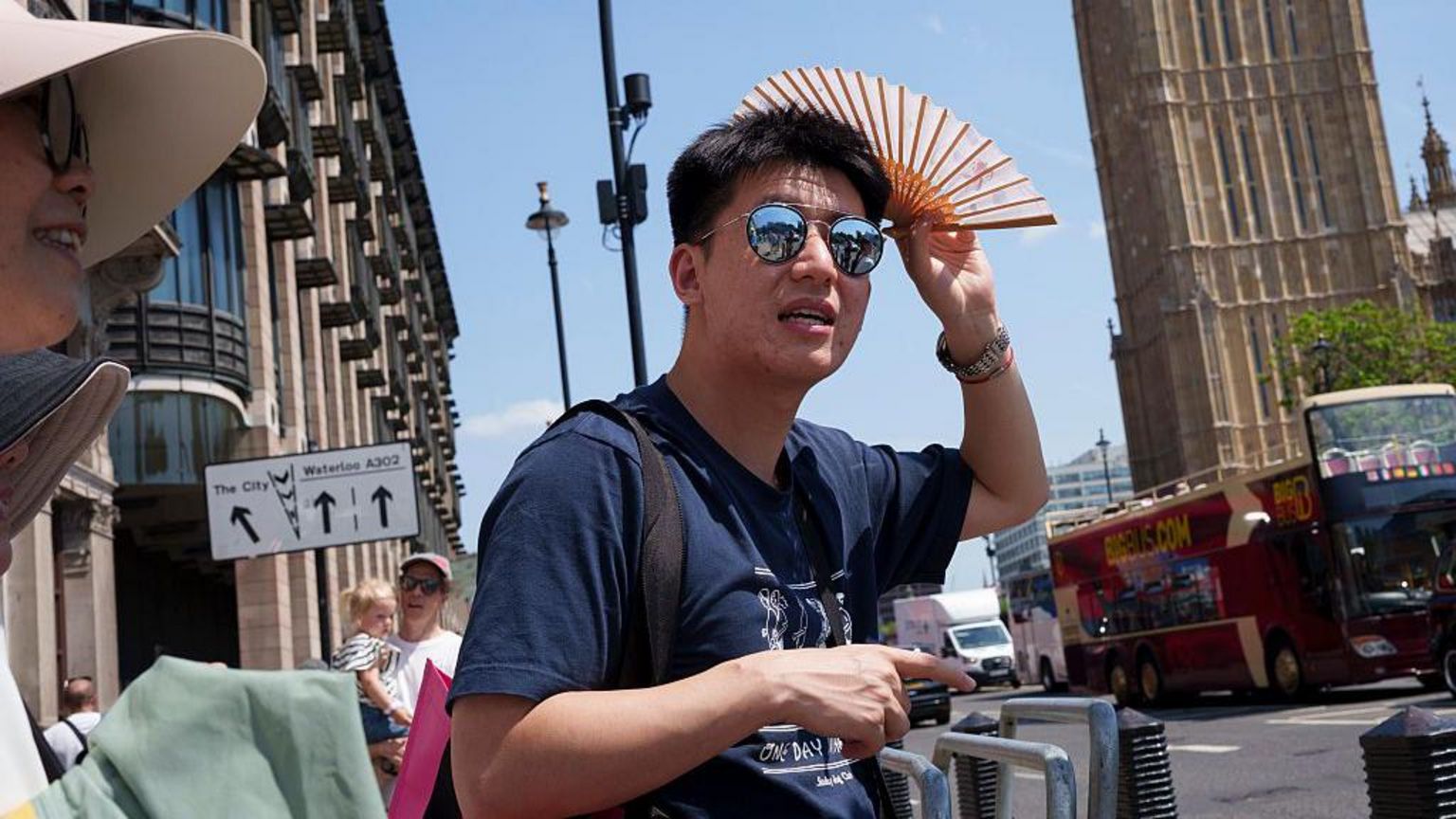 一名男子戴着太阳眼镜，手持摺扇，站在英国伦敦大笨钟前。