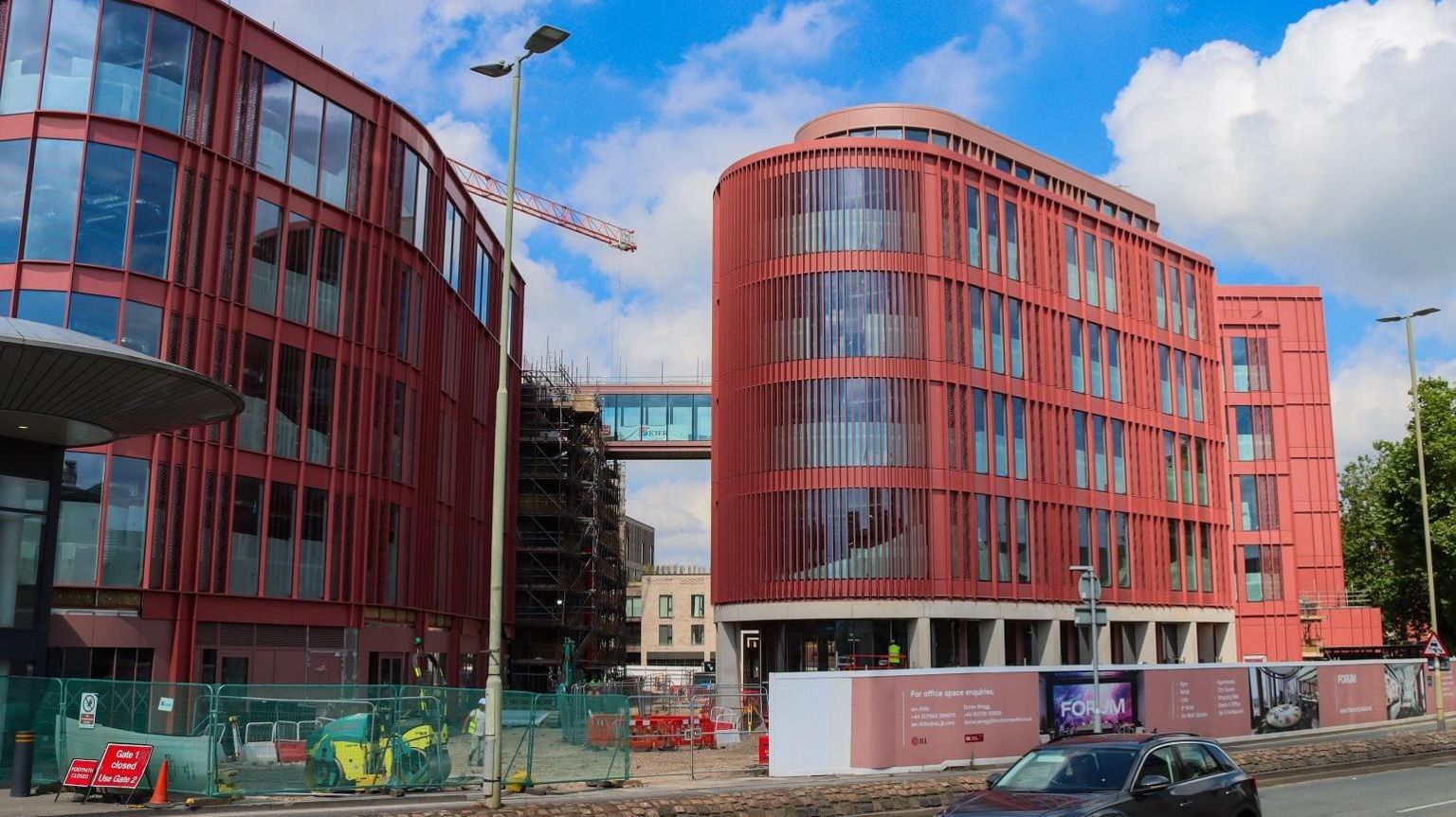 A picture taken from the street of two new buildings. They are both several stories tall and built with distinctive red panelling and curved shapes. 