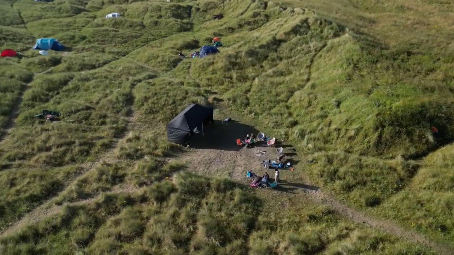 A drone shot of five tents pitched on sand dunes which are covered in grass.