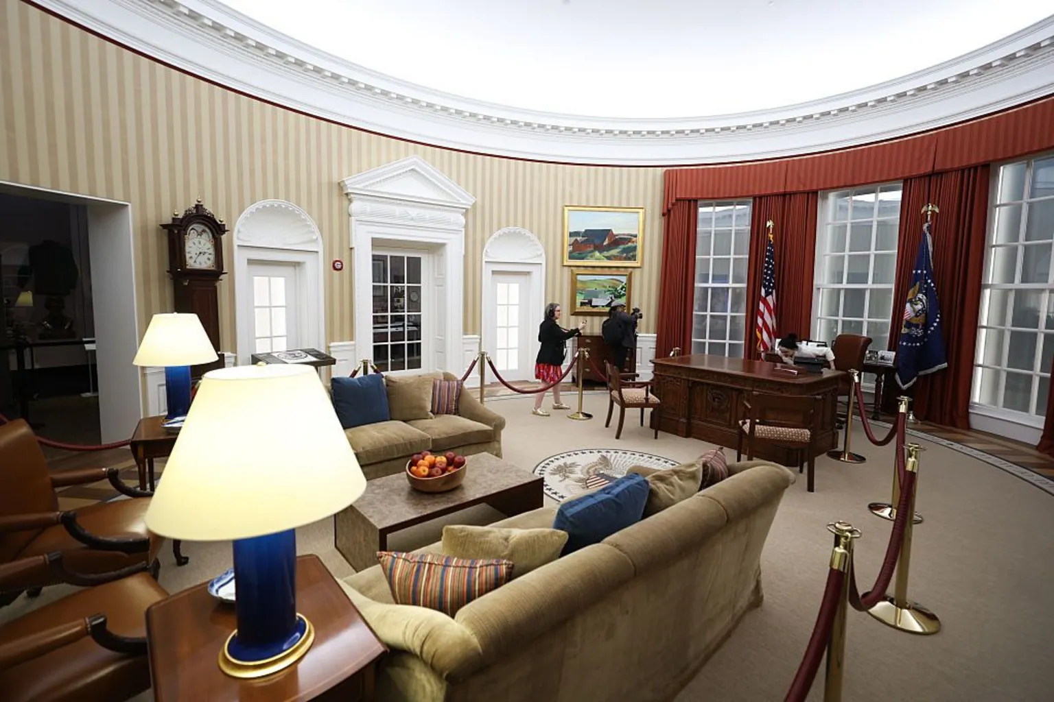 Talia Sprague/Bloomberg via Getty Images Stuffed chairs, lamps and dark wood furniture fill a replica of the Oval Office, with parts of the space sectioned off by red velvet ropes. Two people example
