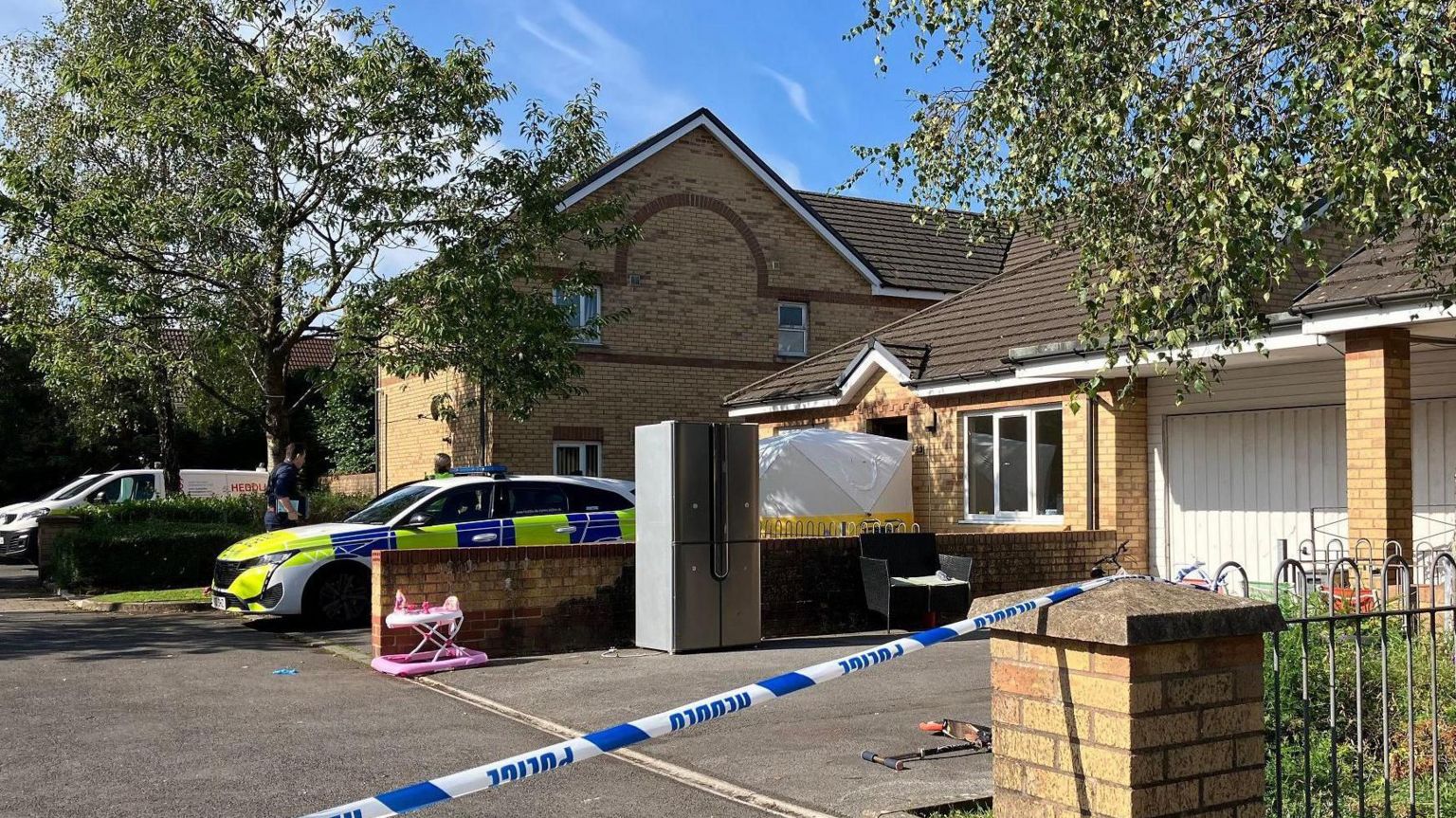 A police cordon outside a property on Cwm Du Close in the Gendros area of Swansea. A pink child stroller is visible behind the police cordon and there is a scooter strewn on the floor. 