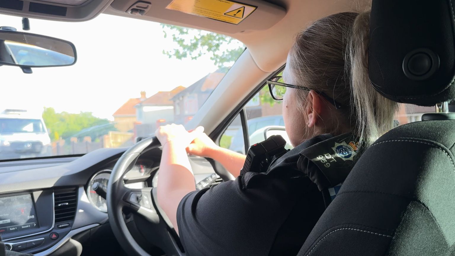 A Sussex police officer is driving a police car, as seen from the rear seat of the car