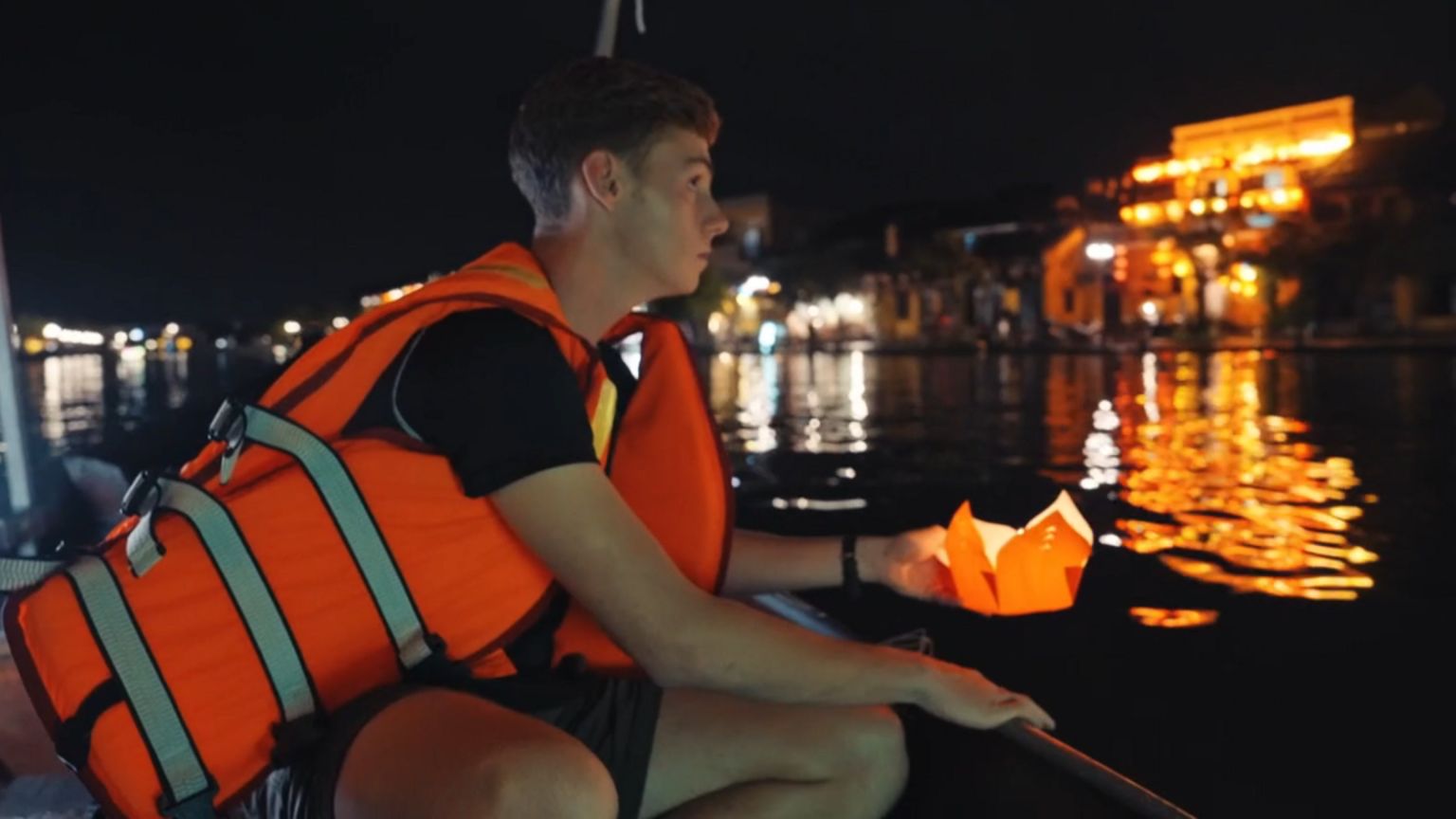 Alfie Watts, wearing a life jacket, leans out of a boat as he prepares to place a lantern with a candle in it on the water