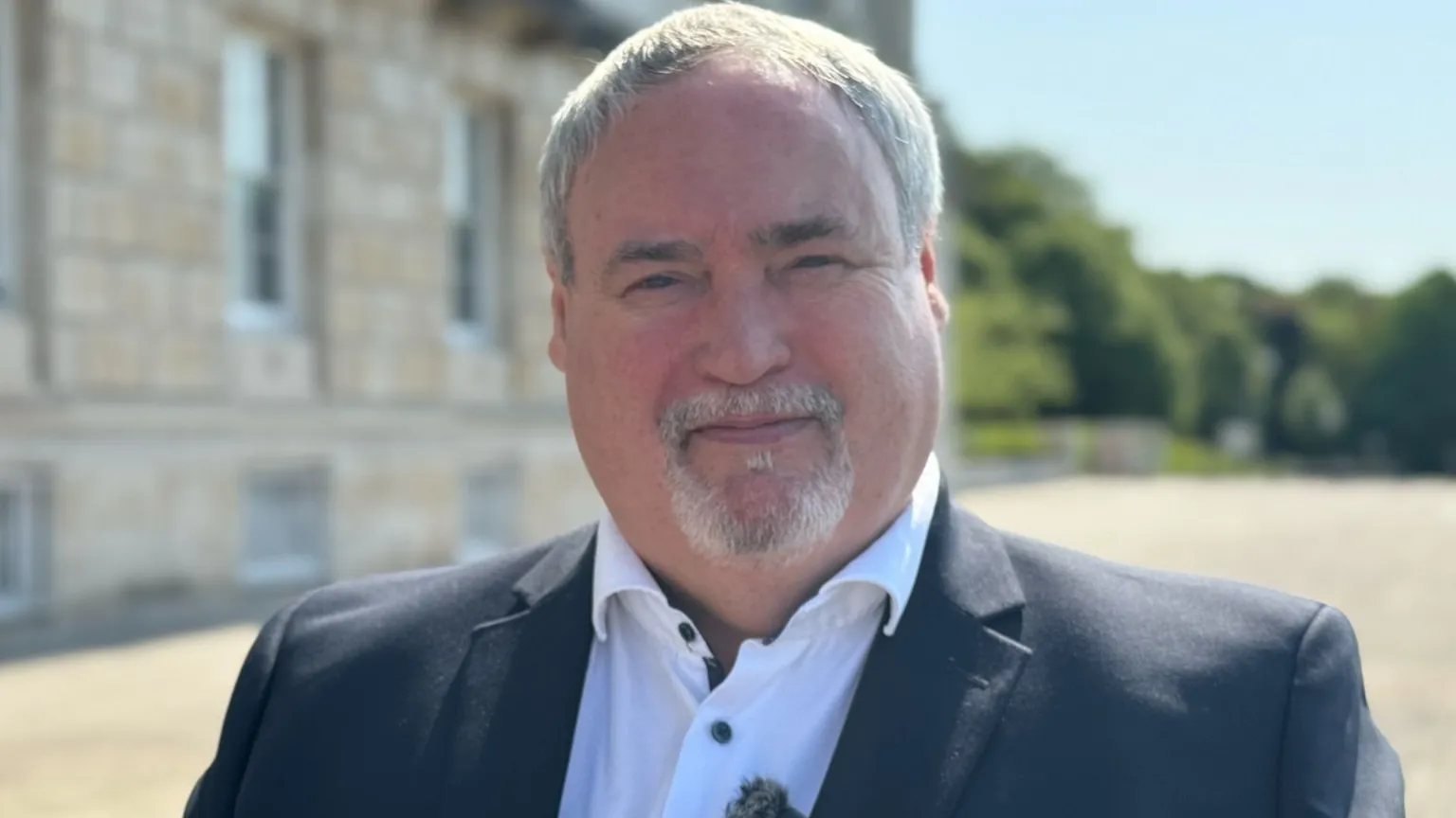 A man with grey hair and beard, he has a dark blazer and white shirt
