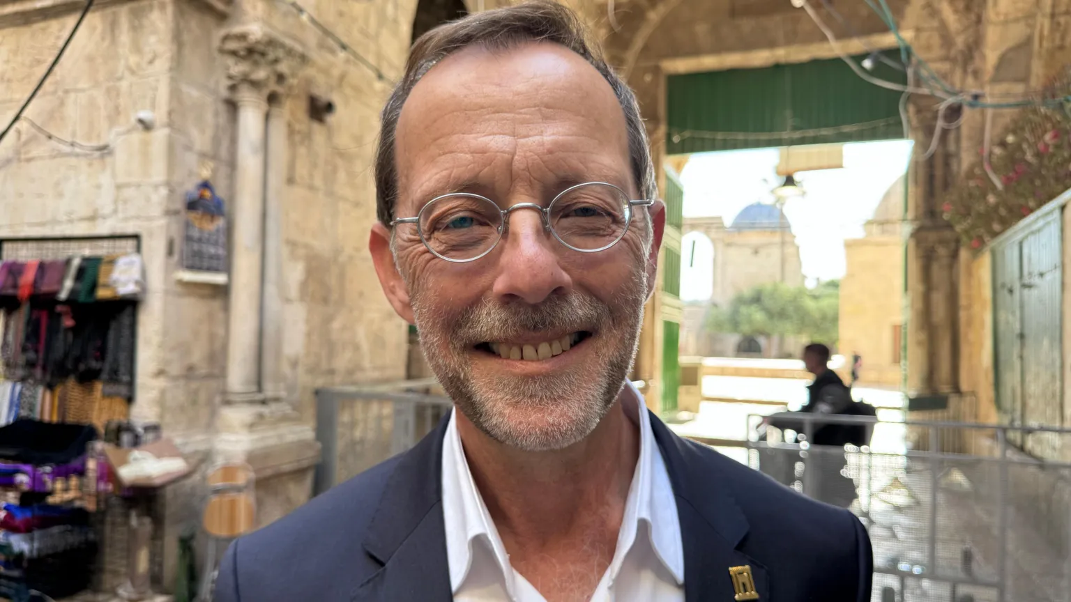 Maarten Lernout/BBC Man with small round metal framed glasses and a grey suit with a weight shirt and a little gold badge on his lapel. He is smiling in the background there is a barrier