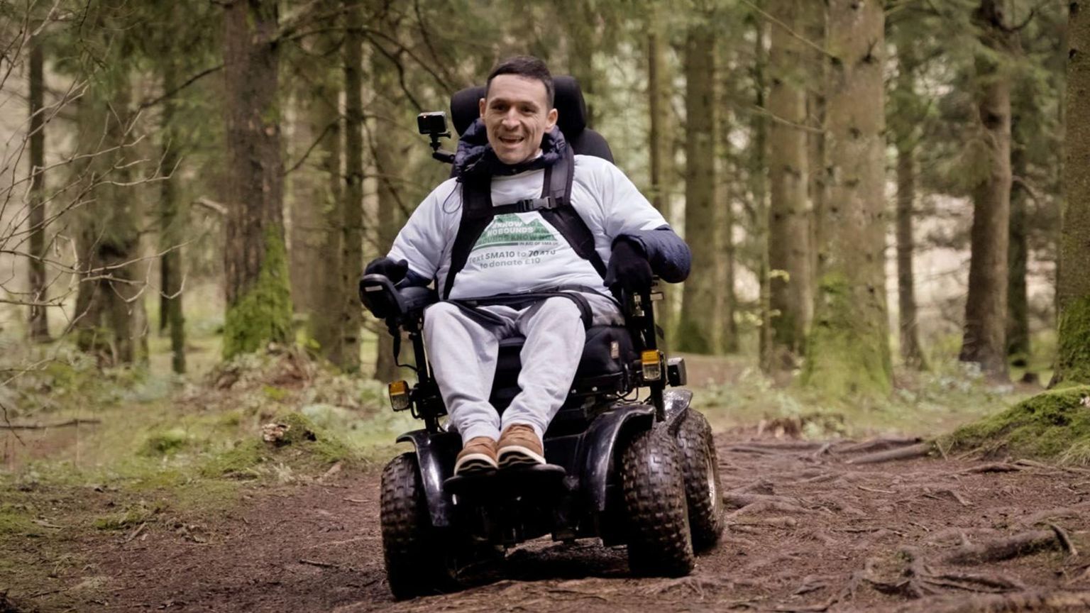 Josh Wintersgill wearing a full grey tracksuit and black gloves. He is smiling as he explores through a forest