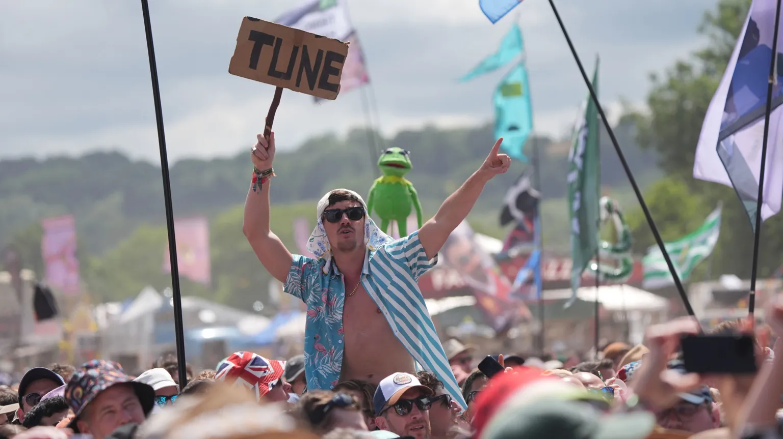 PA Media A man in an open blue and white shirt sits on the shoulders of a friend at Glastonbury festival with a sign that says "Tune". Kermit the Frog is held aloft behind him