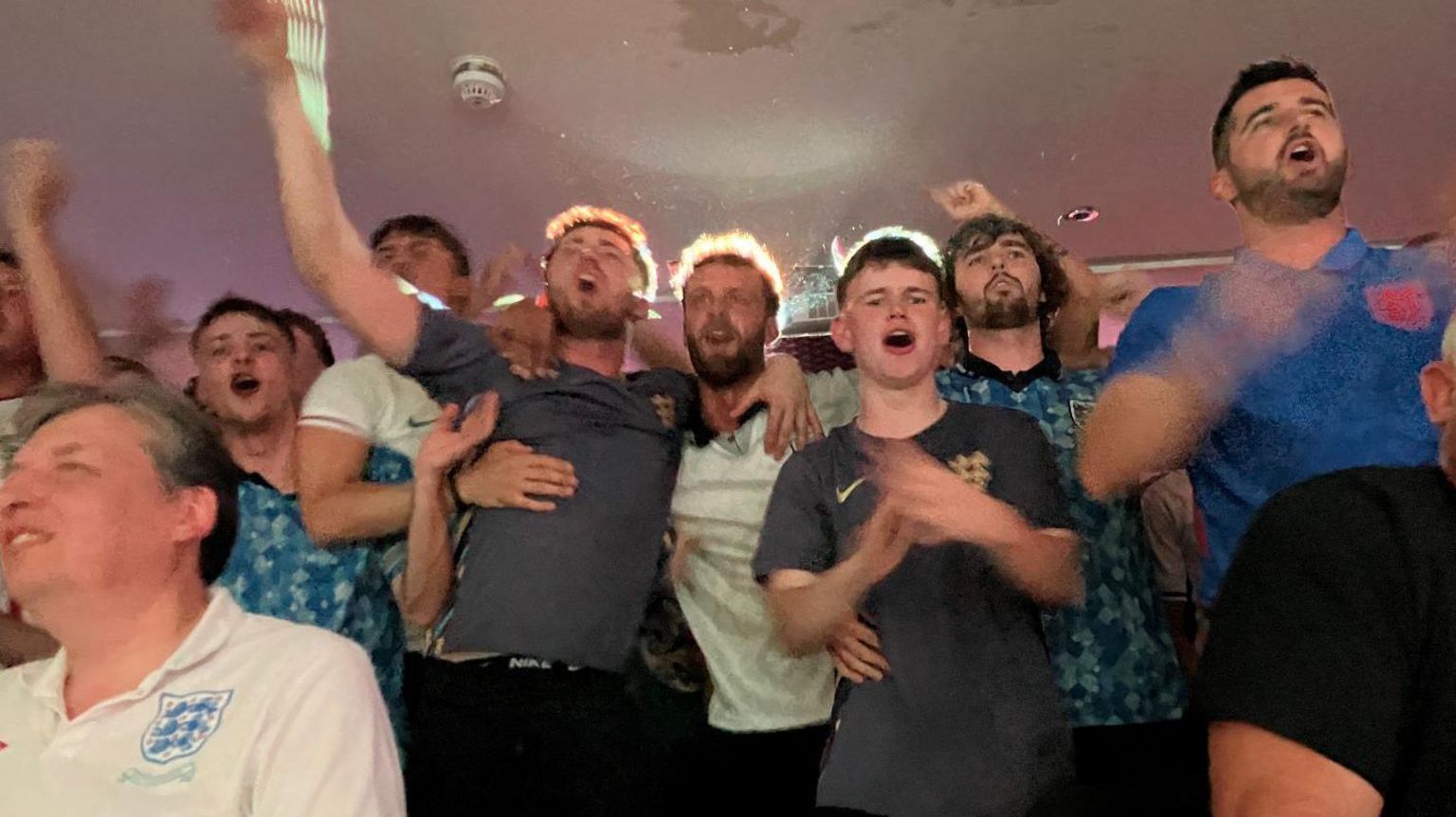 A group of football fans cheer in a pub, with their arms raised