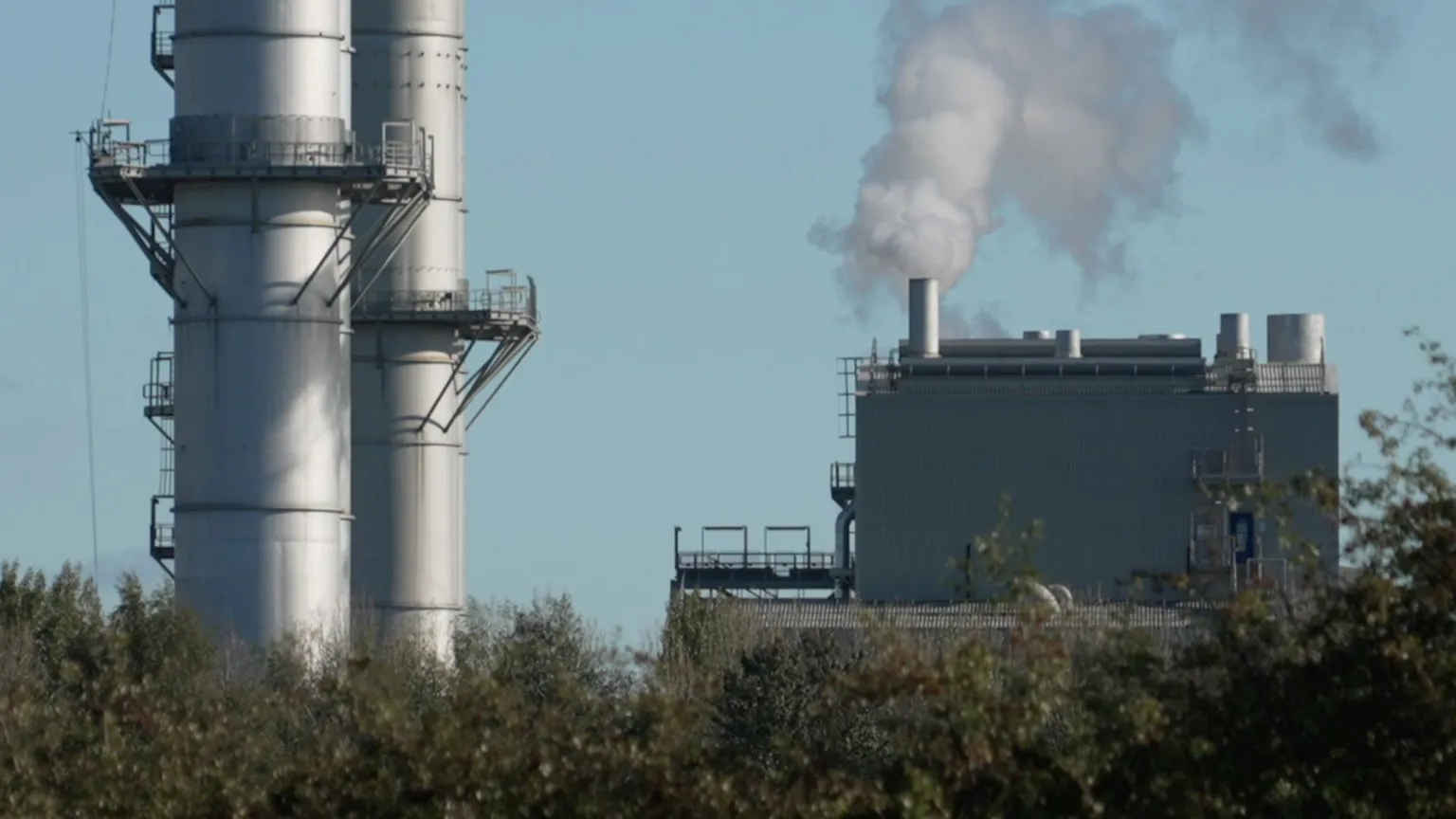 The image shows an industrial power station with large cylindrical structures and smokestacks emitting white smoke or steam against a blue sky. The facility has metal walkways, platforms, and safety railings. Dense vegetation and trees are visible in the foreground, partially obscuring the view of the industrial complex.