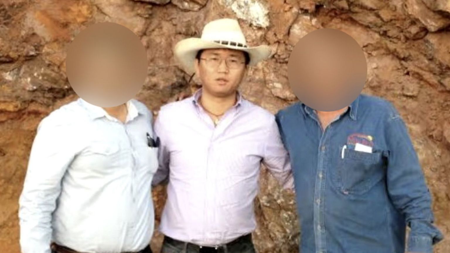 一名男子戴着白色牛仔帽，身穿浅紫色衬衫并将衣襬束进裤内。他戴着眼镜和项鍊，正望向镜头。他左侧及右侧各有一名男子，这两人的面容均经模糊处理，以隐藏其身份。