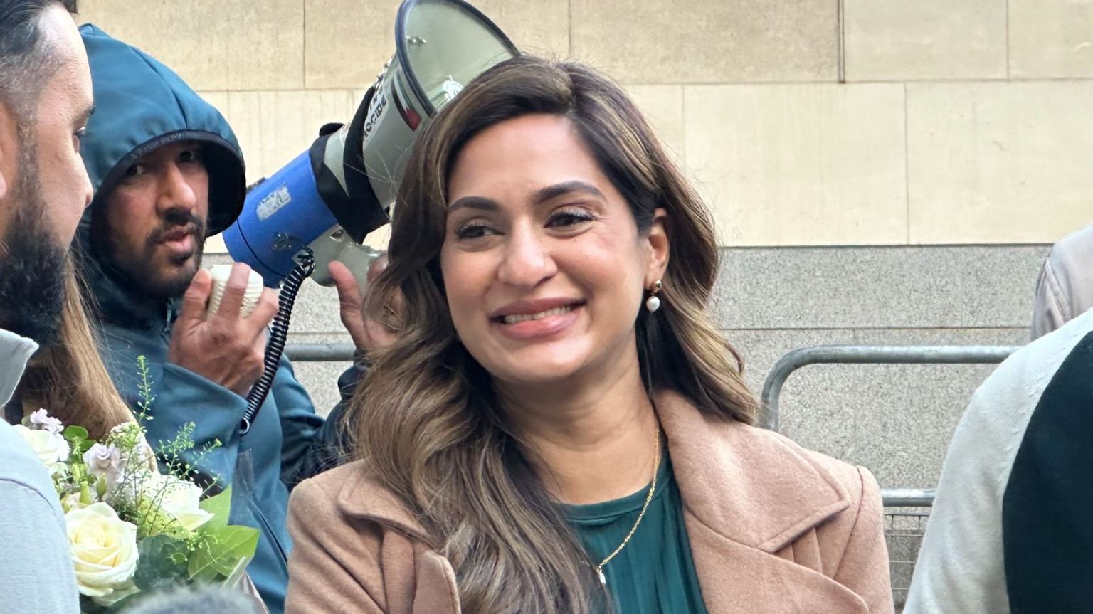 Marieha Hussain, standing outside the courtroom, smiling. She's wearing a beige winter jacket and a blue-green top. 