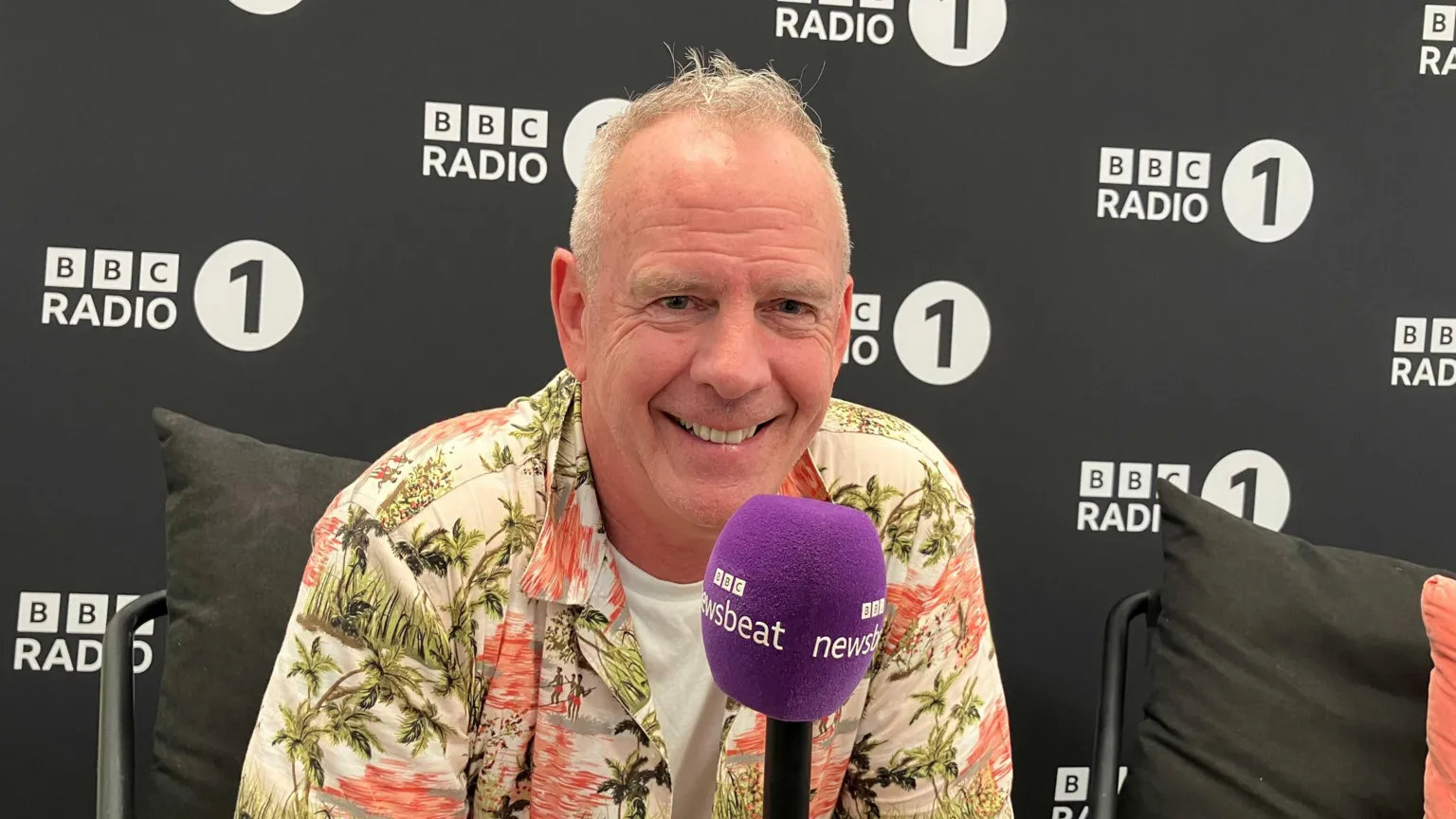 Fatboy Slim smiles holding a purpose Newsbeat microphone. He is wearing an unbuttoned Hawaii shirt over a white t-shirt. He is in front a black backdrop, with the Radio 1 logo on.