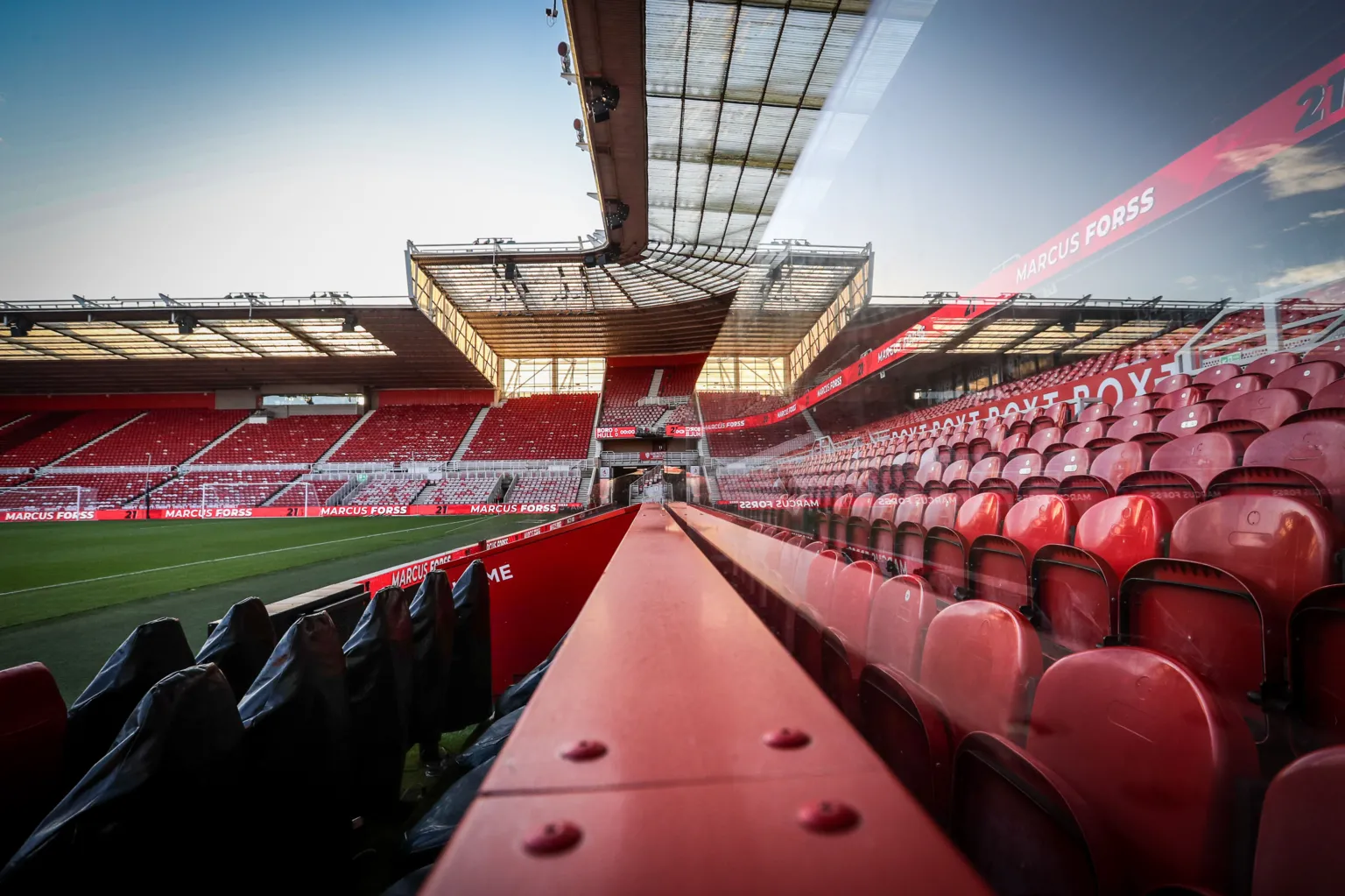 Tom Banks/MFC An empty Riverside Stadium. The shot is taken along a steel barrier behind the dug-outs. Rows of red seats and the glass front of a line of executive boxes can be seen on the right. In the distance is the two-tiered stand behind the goal.