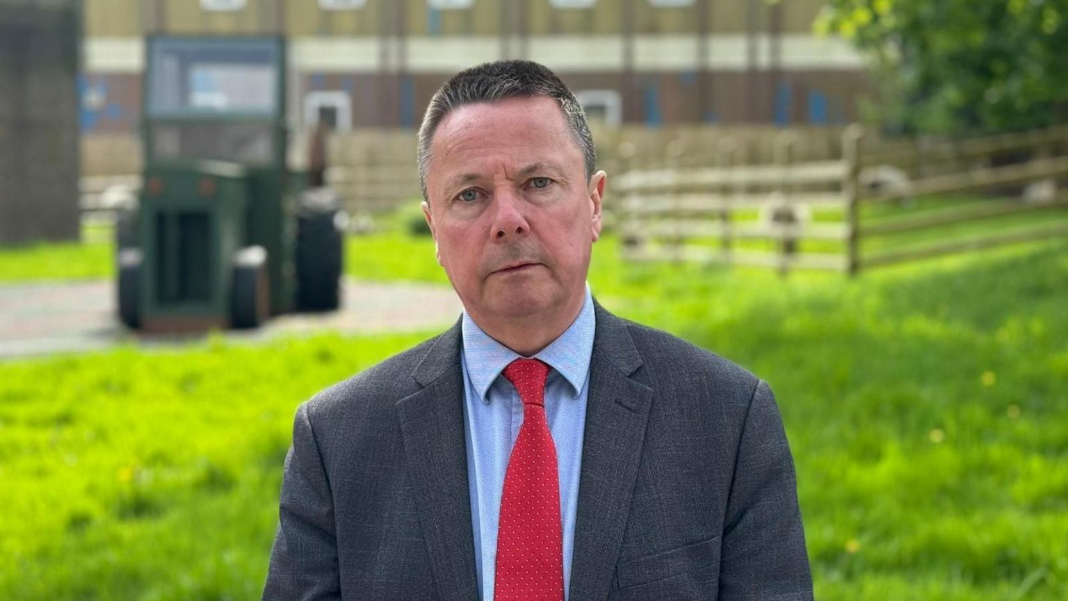 Man standing in a paddock looking into the camera. there is an out-of-focus camera behind him. He's wearing a grey suit, red tie and blue shirt.