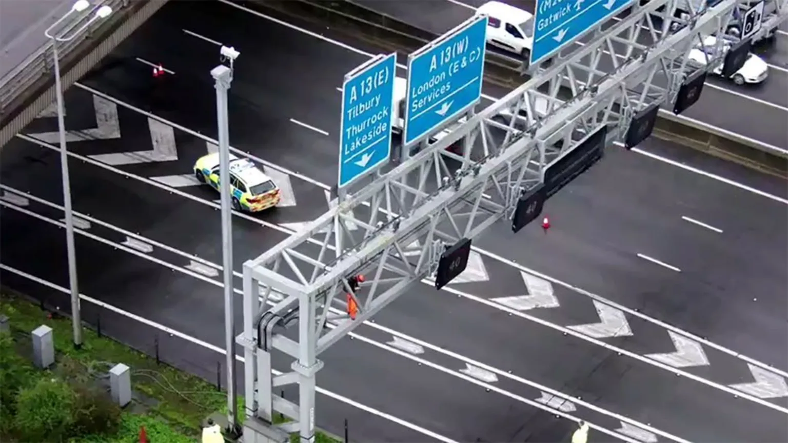 An aerial view shows a collapsed overhead gantry structure blocking multiple lanes of a major highway. The white metal framework has fallen across the road surface, with blue highway signs visible. A police vehicle with high-visibility markings is positioned nearby, and traffic lanes are clearly marked on the asphalt.