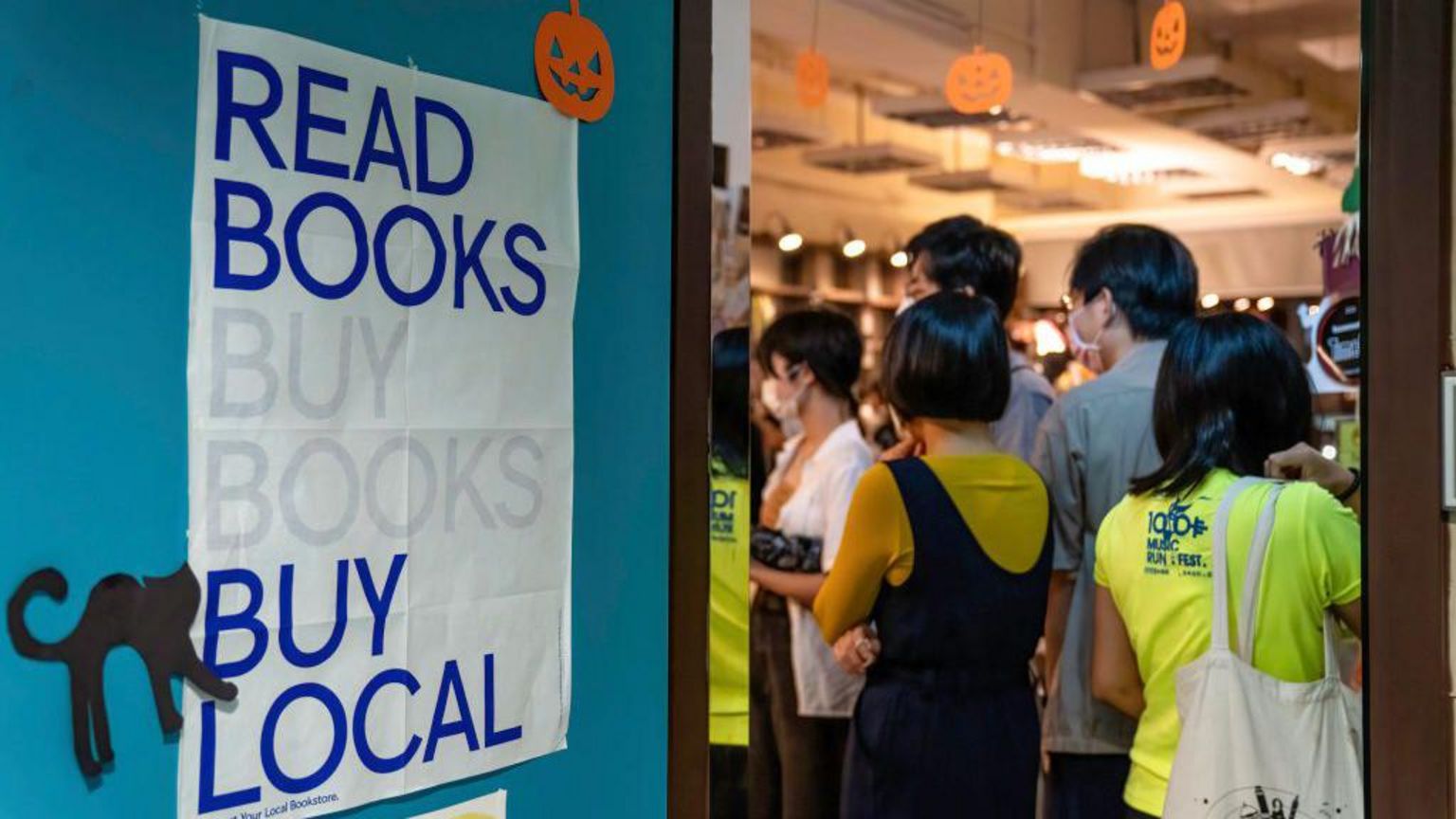 顾客正在浏览书籍，店门上贴着“阅读、购书、支持本土书店”（Read Books, Buy Books, Buy Local）的海报。