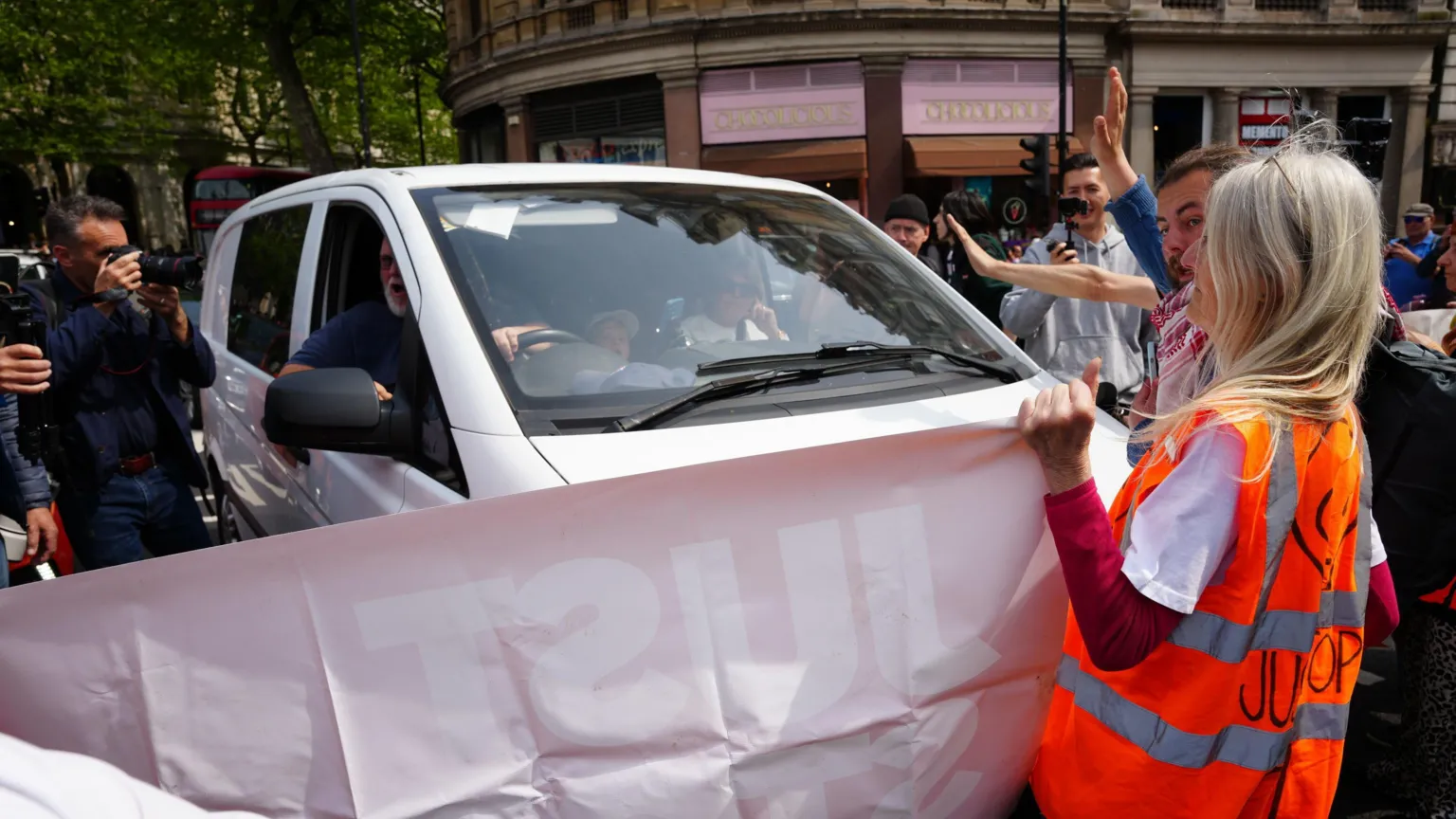 The image shows environmental protesters holding a large pink banner in front of a white minivan on a street. A woman in a high-visibility orange vest is positioned near the vehicle's front. Onlookers and photographers surround the scene, capturing this disruptive protest action.