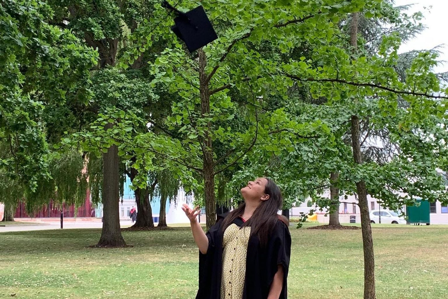 Abbie Tutt in clothing worn for graduation, throwing her hat in the air in front of a park with a tree directly behind her.