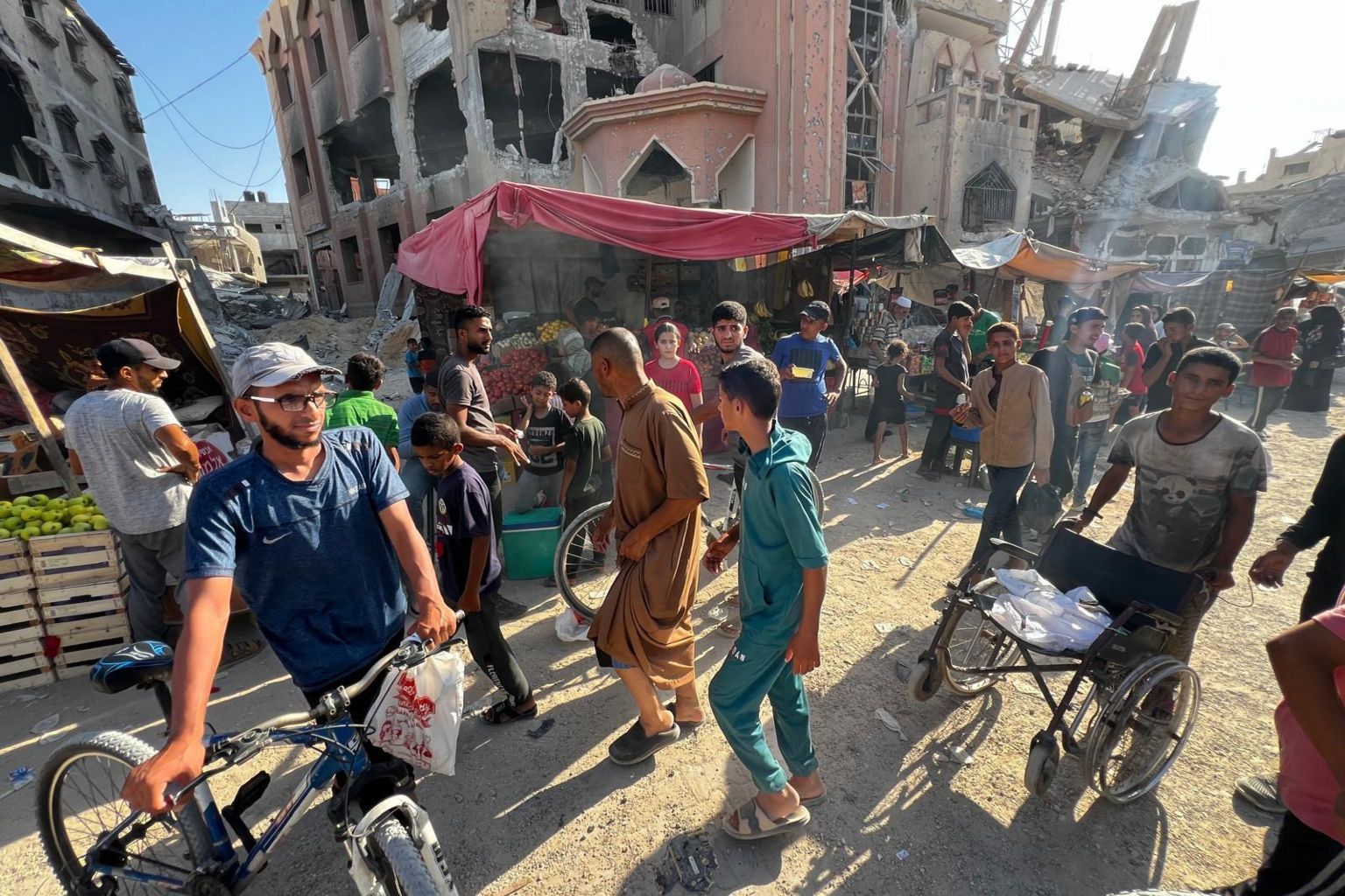 People are seen in front of market stalls in Khan Younis, with destroyed buildings behind them