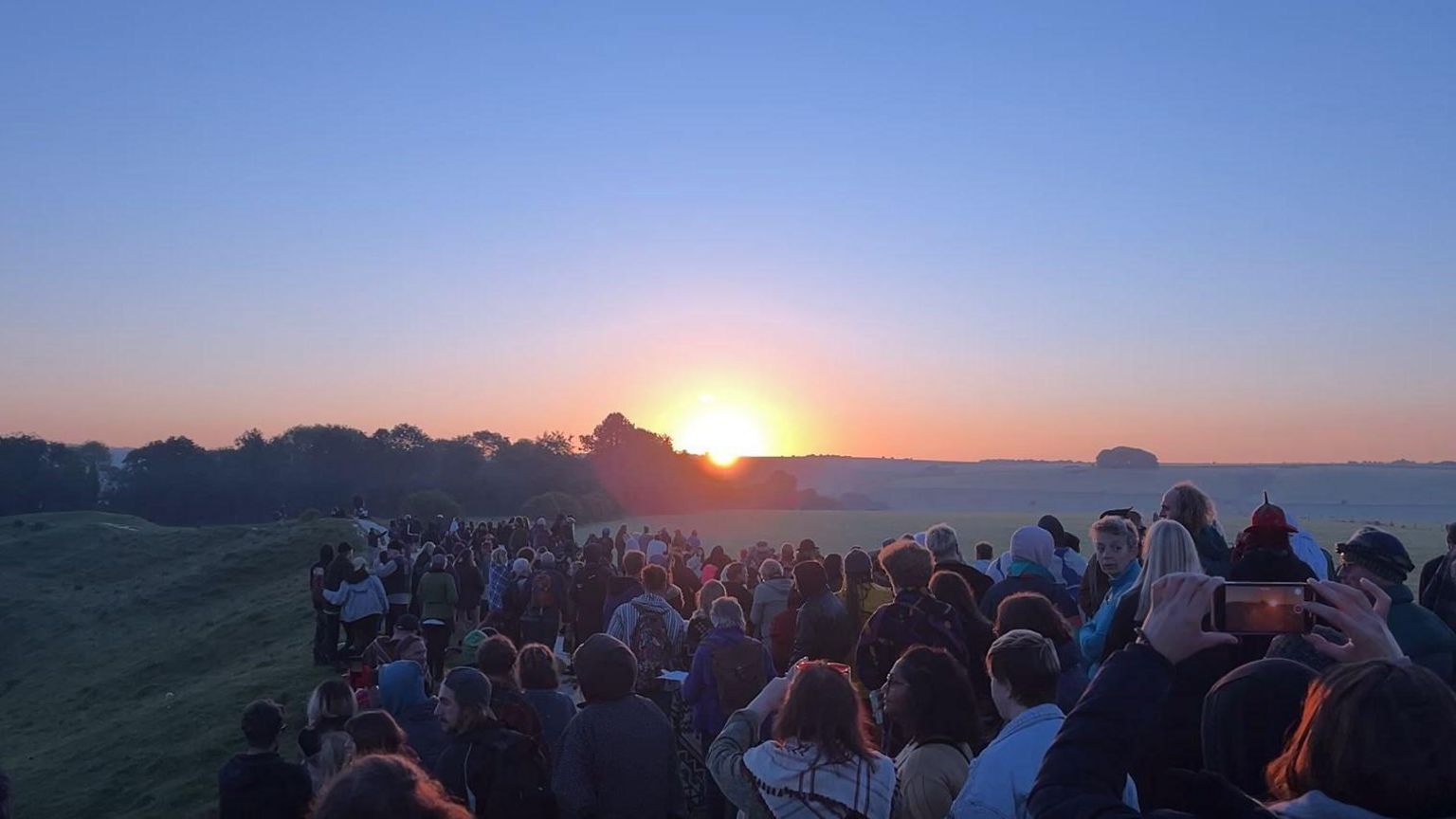 Crowd of people gathered along a bank at Avebury, with the sun rising