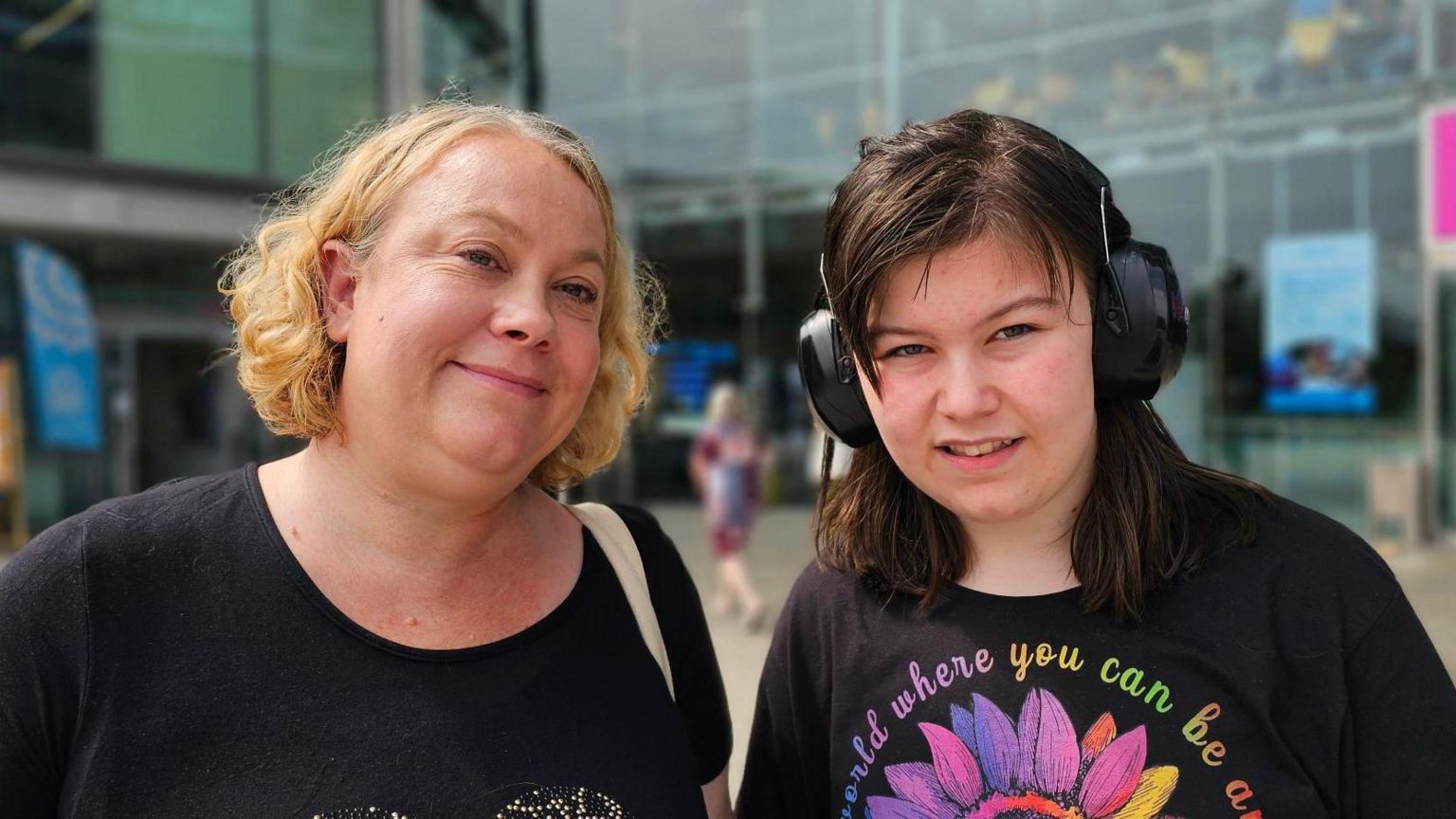 A closed mouth smiling Victoria Trattles who has blonde chin-length hair, wearing a black t-shirt with her daughter Maria who has dark shoulder-length hair and is wearing ear protectors and a black t-shite with a multi-coloured flower on it 