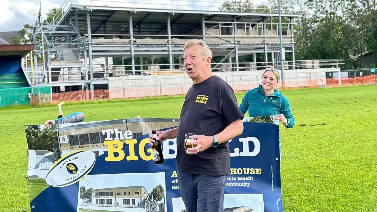 Alan Dunn raising a glass on the pitch, in front of the metal frame of the new building. He stands in front of a sign saying 'The Big Build'. A girl with blonde hair and a green top can be seem in the background