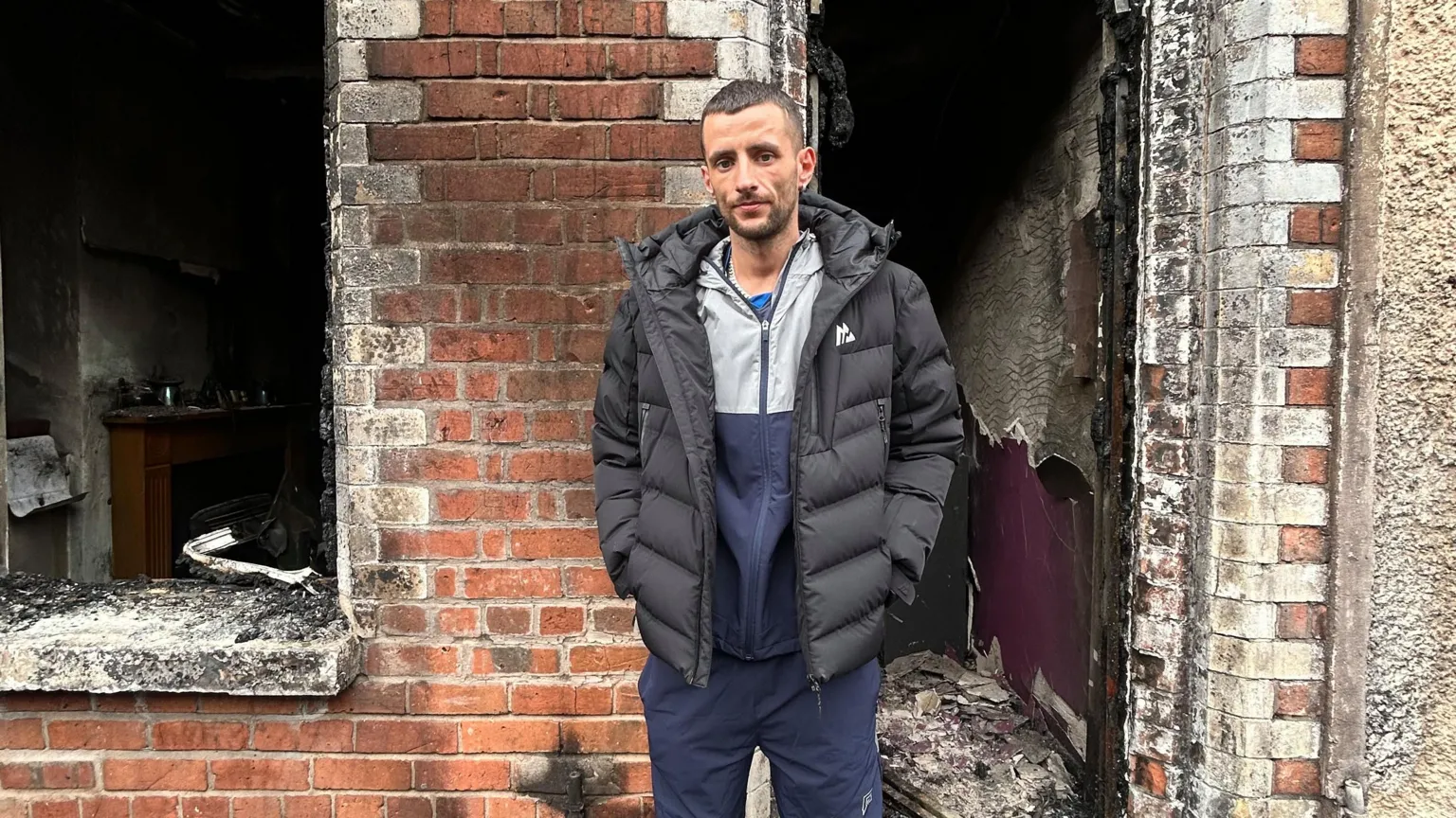 BBC A man with brown hair and a black puffer coat stands outside his home