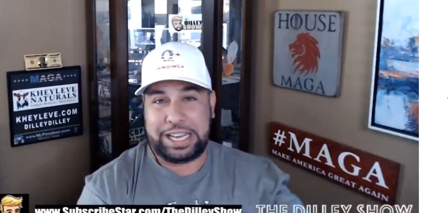 Brenden Dilley seen in one of his online videos, wearing a white baseball cap and staring at the camera with a big "MAGA" sign on the wall next to him 